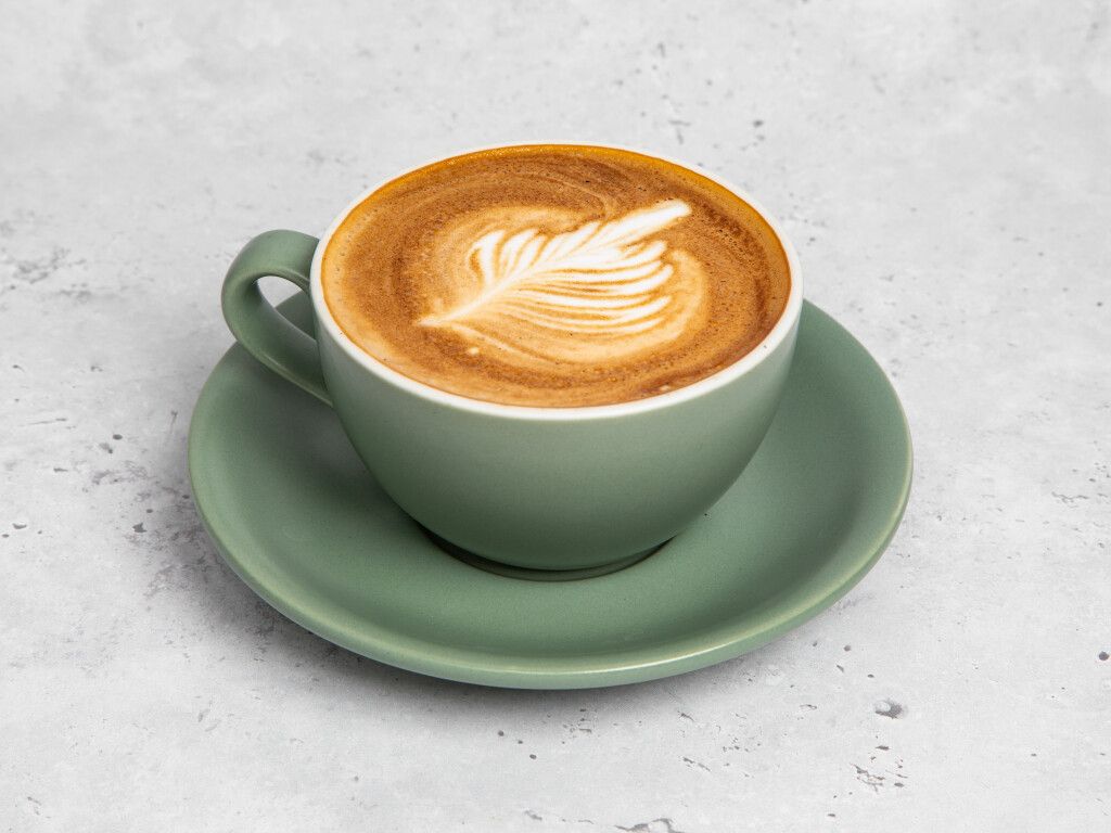A latte with latte art in a sage green mug on a matching saucer, set against a gray background.