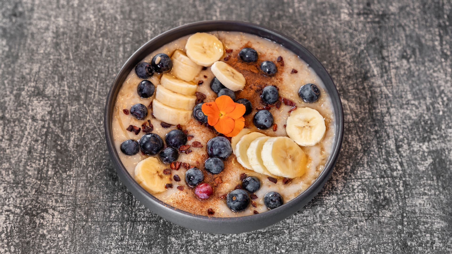 Oatmeal bowl topped with sliced bananas, blueberries, cocoa nibs, and an orange flower, on a gray background.
