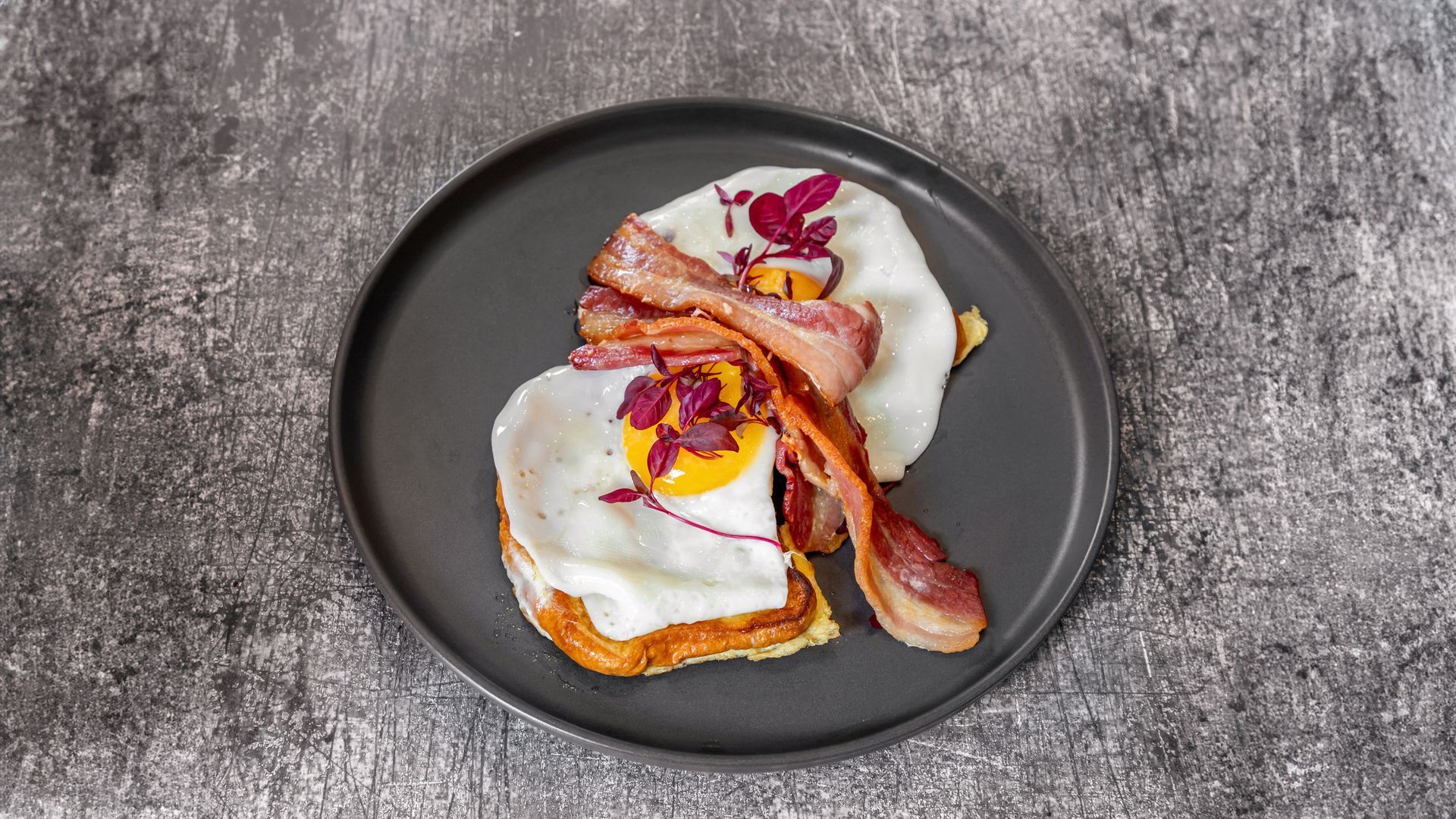 Fried eggs, bacon, and greens on toast, on a gray plate, set on a gray surface.