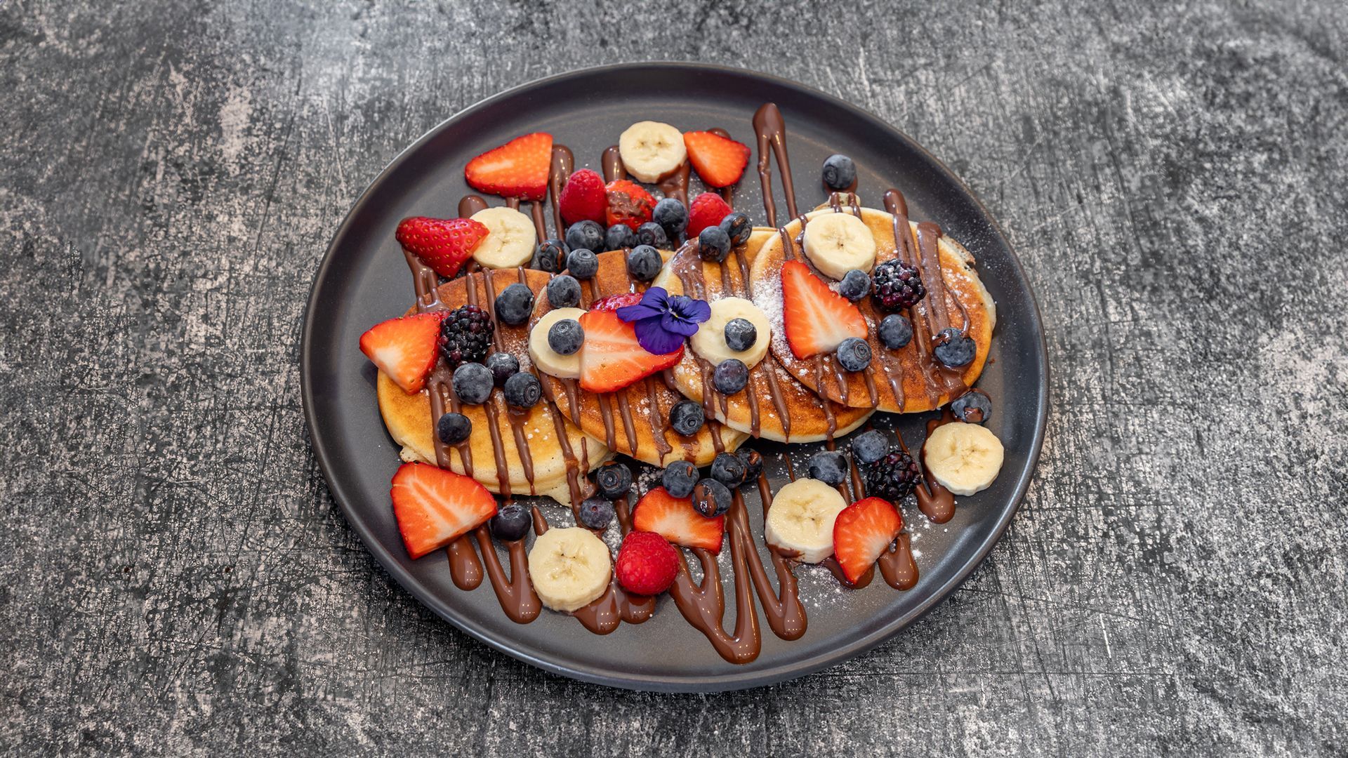 Pancakes topped with strawberries, blueberries, banana slices, and chocolate drizzle on a dark plate.