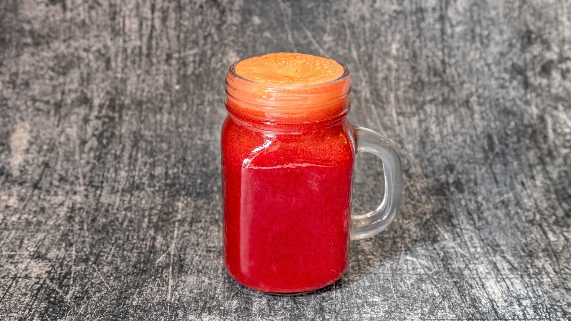 Red juice in a clear glass mug with a frothy top, set against a mottled gray background.