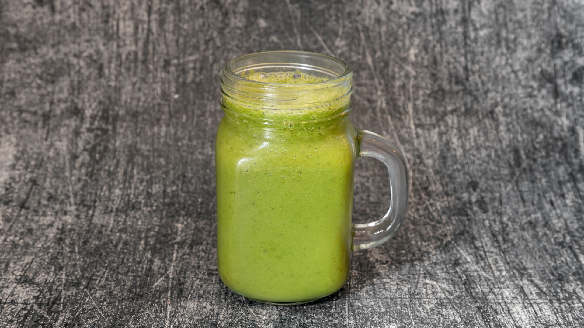 Green smoothie in a clear mason jar on a mottled gray surface.