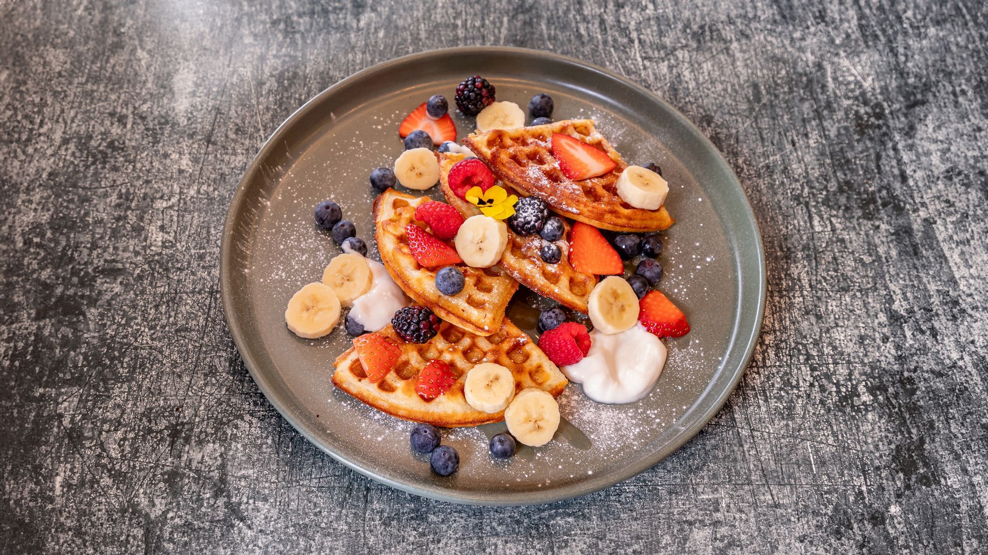 Waffles with strawberries, blueberries, blackberries, banana slices, and cream on a gray plate, dusted with powdered sugar.