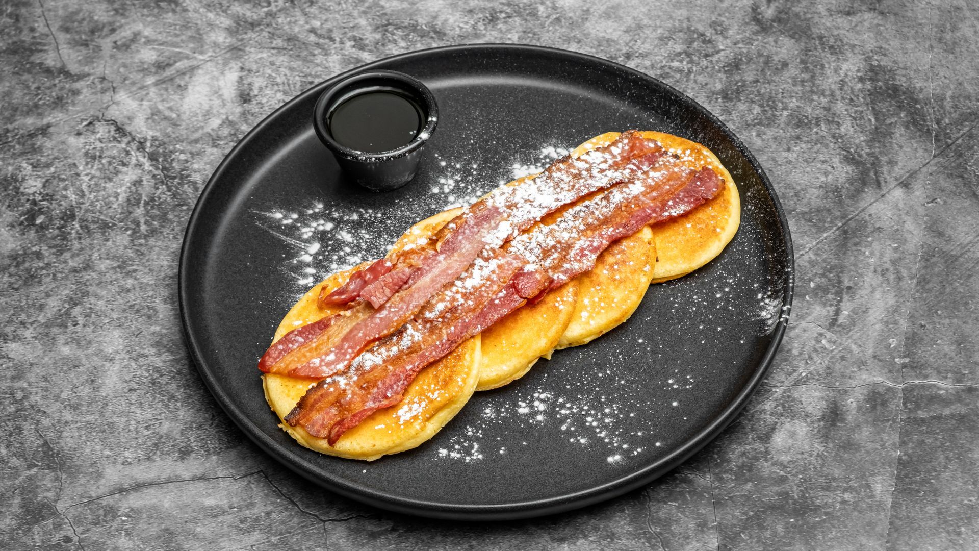Pancakes topped with bacon and powdered sugar, served with syrup on a black plate.