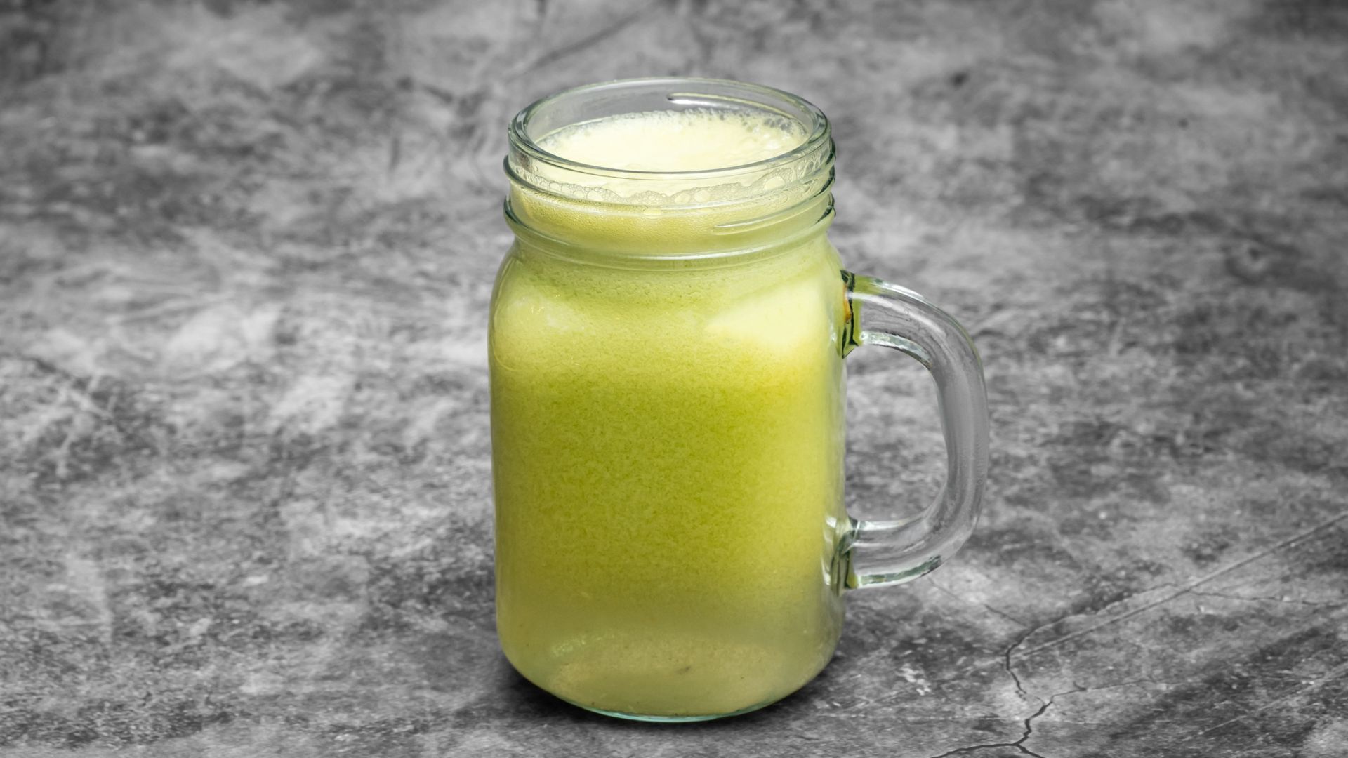 Green smoothie in a mason jar with a handle, against a textured gray background.