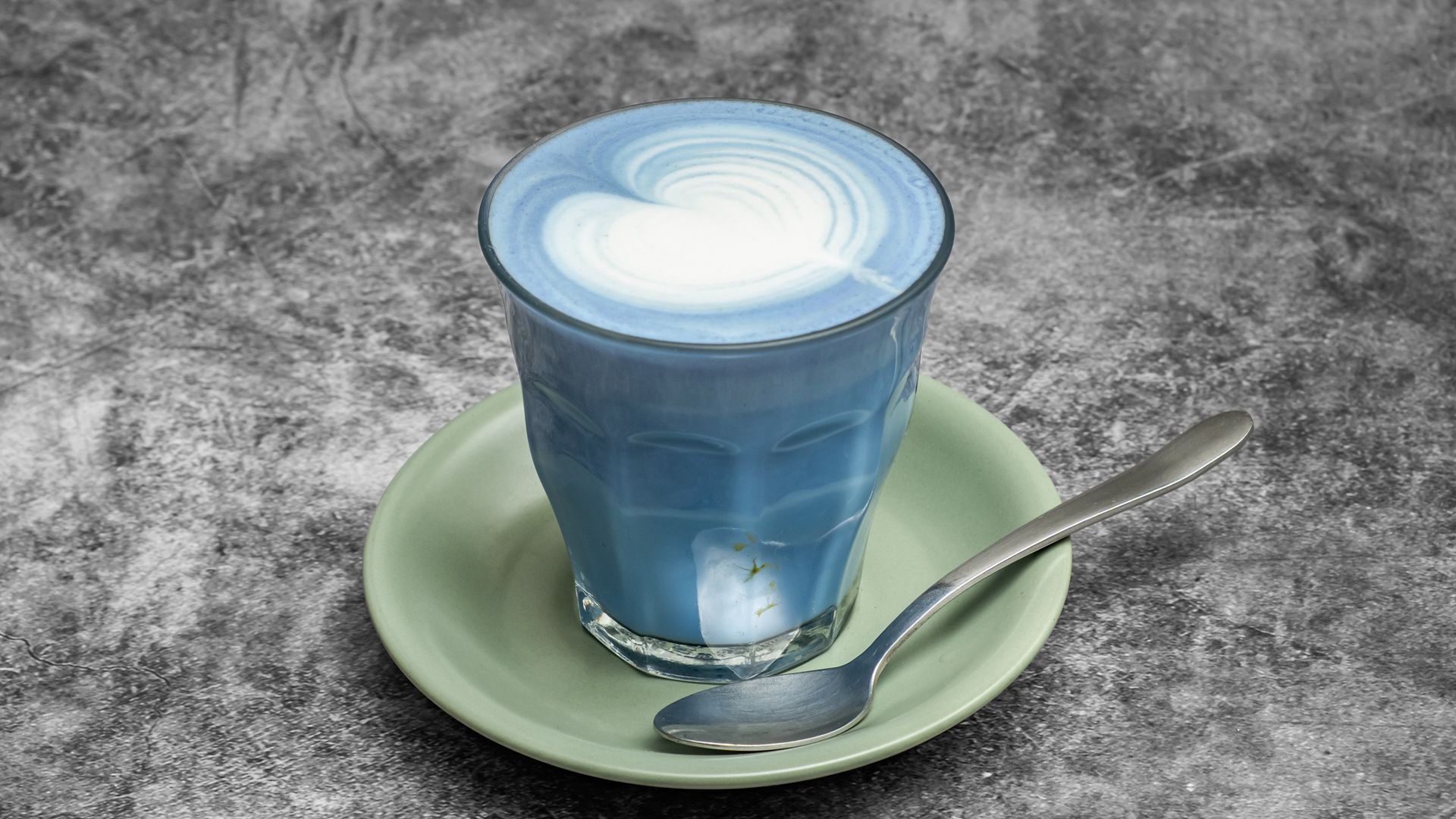 Blue latte in a glass, with a white heart design, on a green saucer with a spoon.