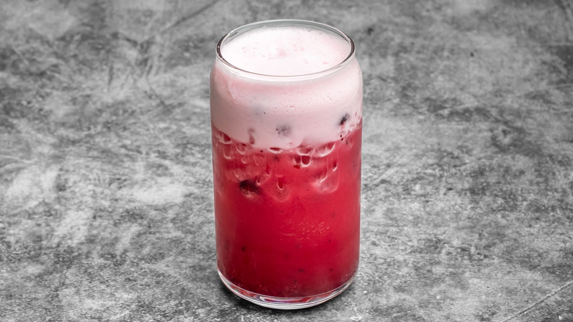 A tall glass of red and pink beverage with ice on a textured gray surface.