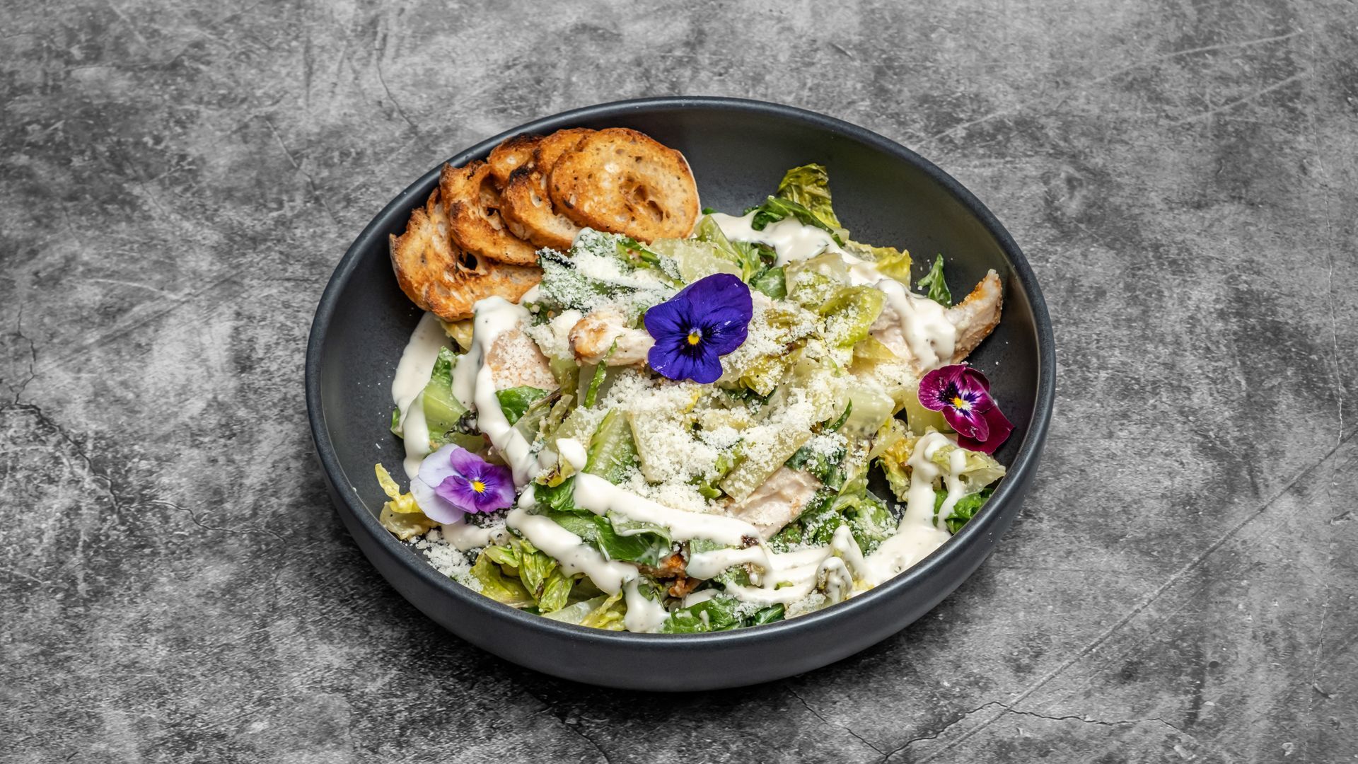 Caesar salad in a dark bowl with croutons, purple edible flowers, and white dressing.