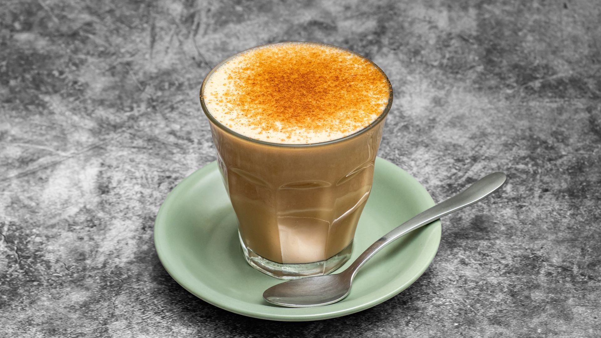 Glass of brown drink on a light green saucer with a spoon; sprinkled with brown powder, set against a gray background.