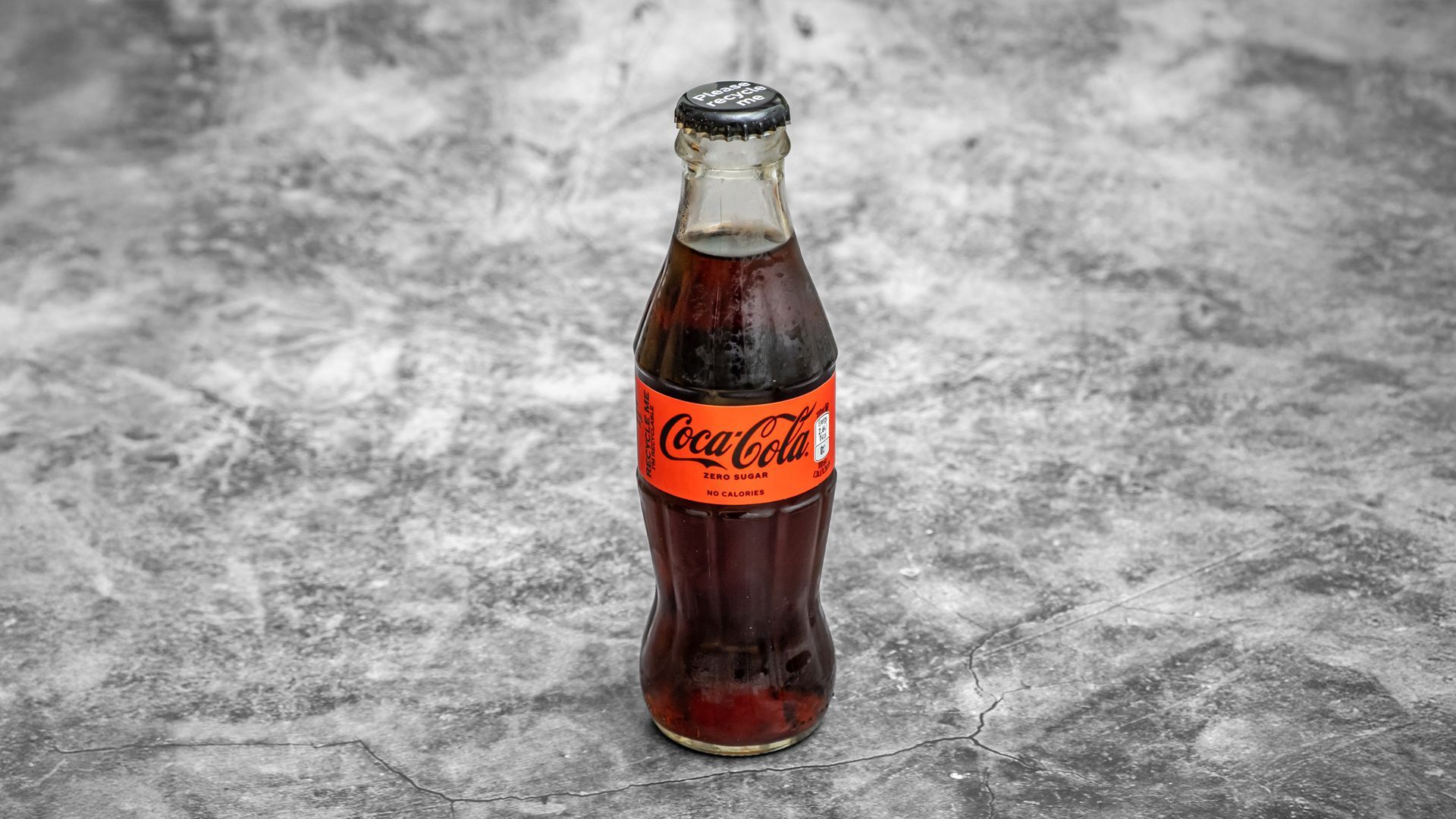 Coca-Cola bottle with red label, cap and dark liquid against a gray background.