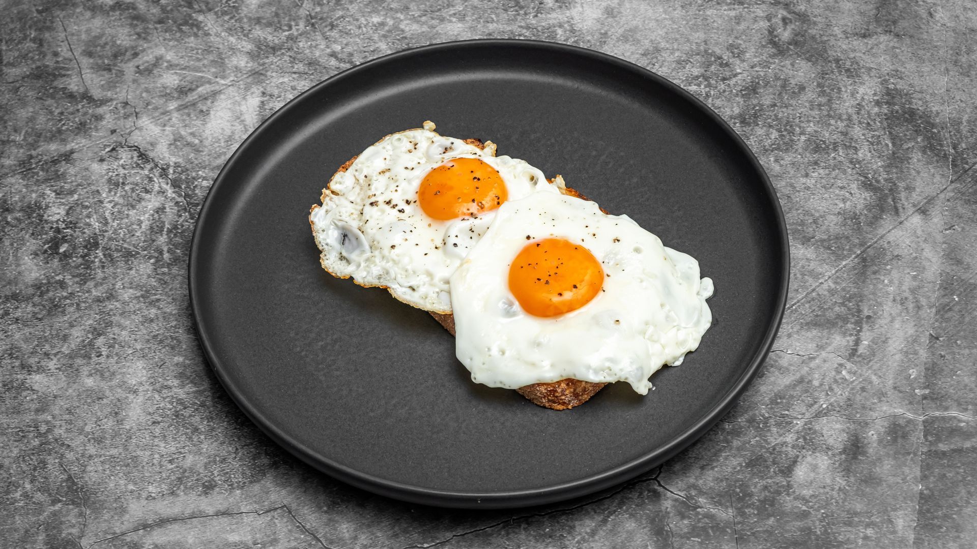 Two sunny-side-up eggs on a slice of toast, seasoned with pepper, on a black plate.