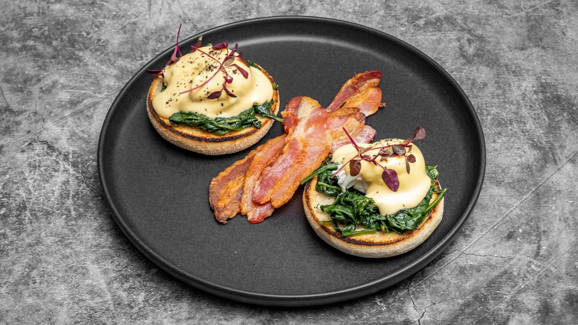 Eggs Benedict with bacon and spinach on a black plate against a gray backdrop.