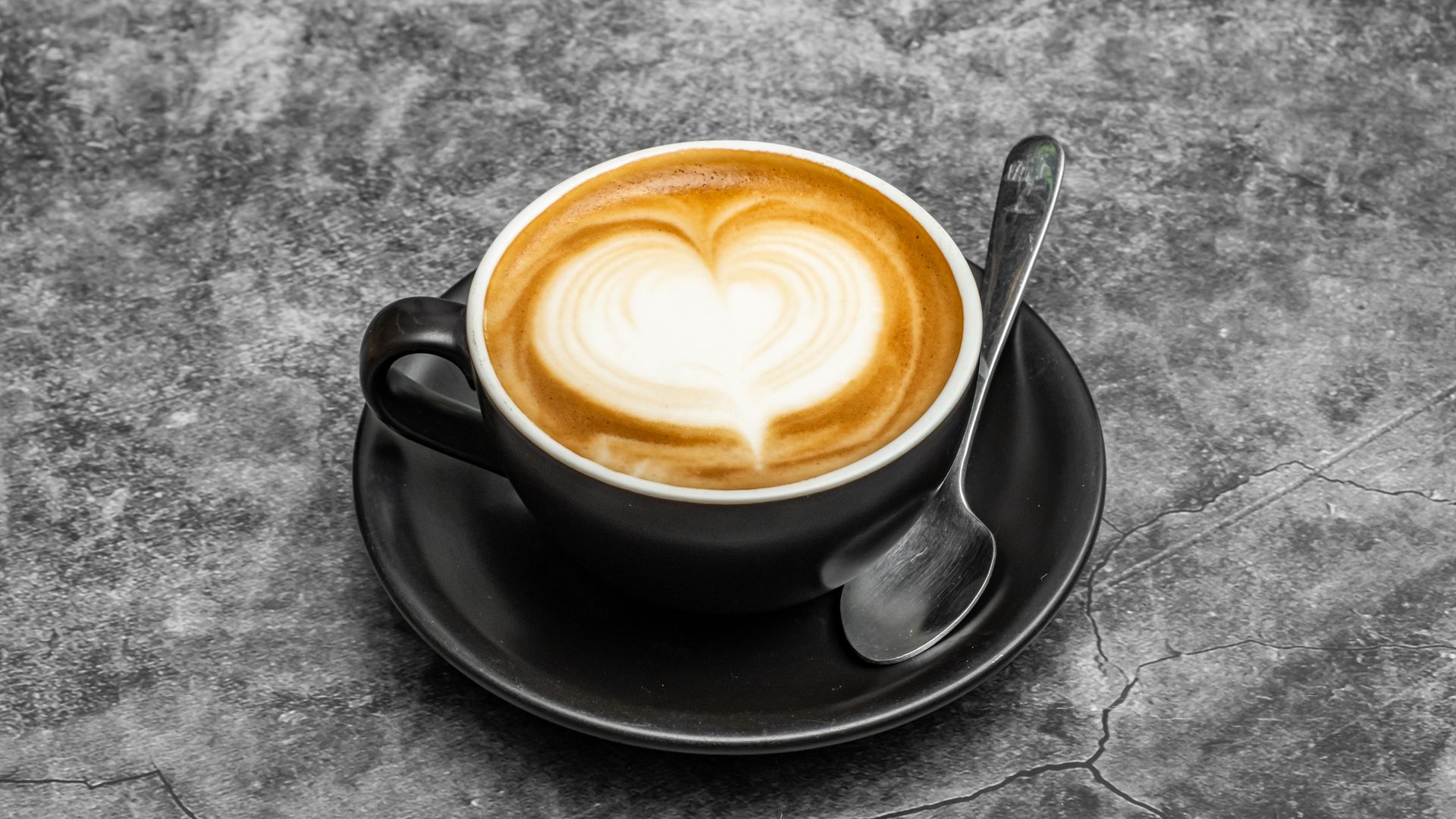 Cup of latte with heart-shaped foam art, on a black saucer with spoon. Gray background.