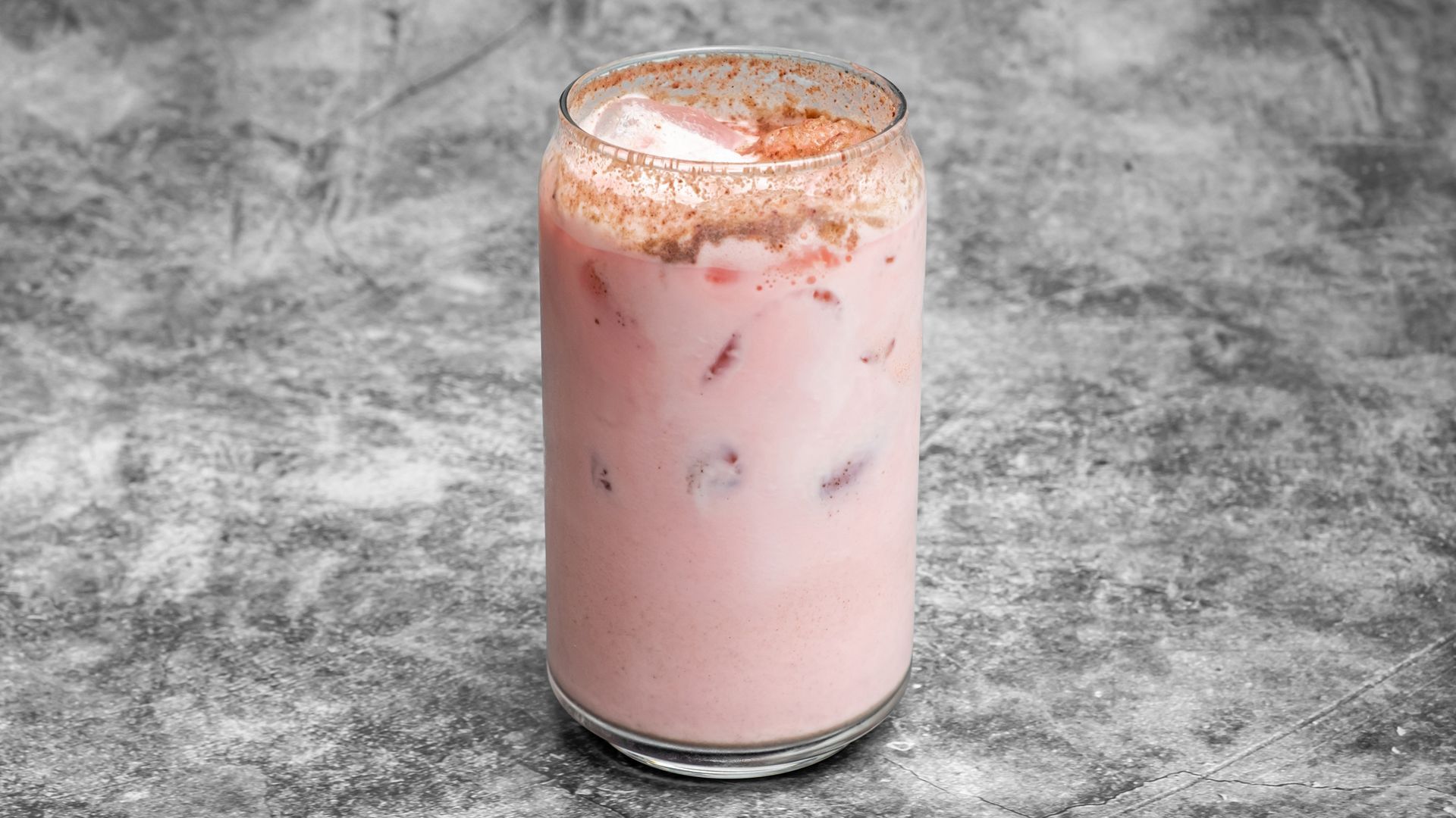 Pink drink in a tall glass with ice, topped with a dusting of cinnamon on a grey background.