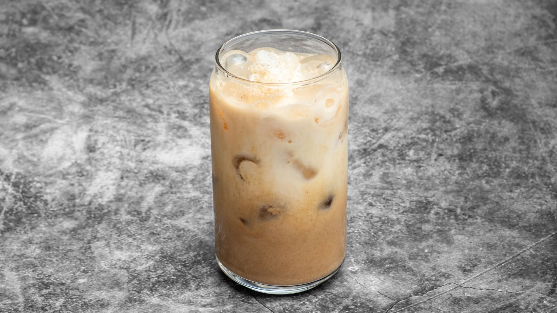 Iced coffee in a clear glass, ice cubes visible, against a mottled gray backdrop.