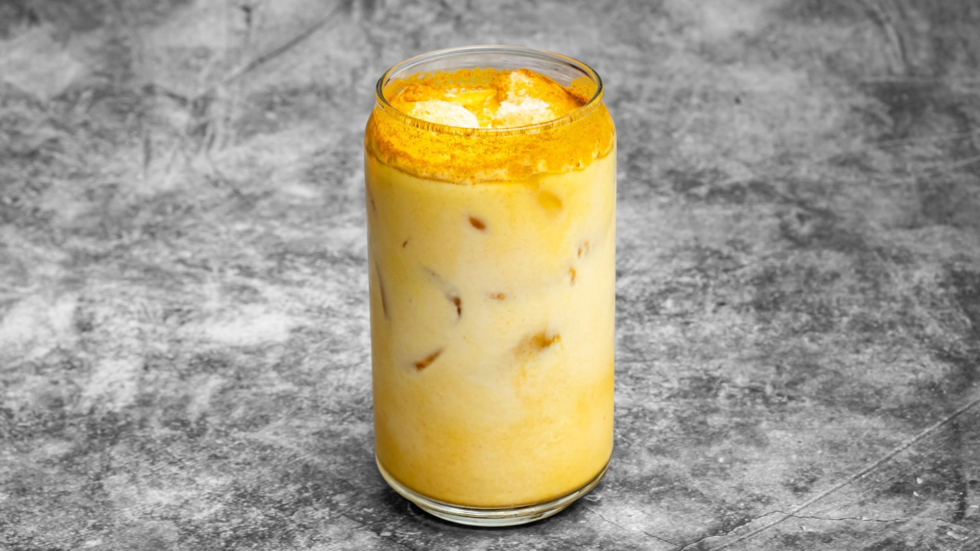Iced drink with yellow rim, beige inside, and ice cubes in a clear glass. Set against a mottled gray backdrop.