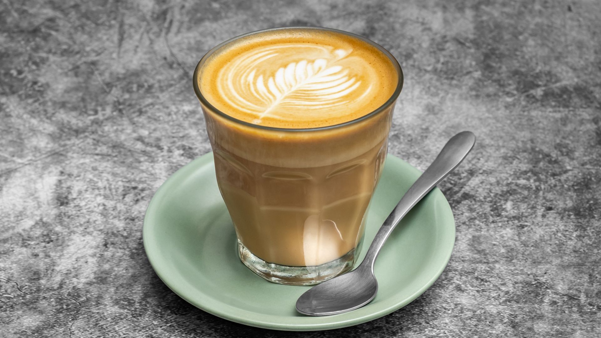 Latte in a glass, with latte art, on a light green saucer with a spoon, against a gray background.