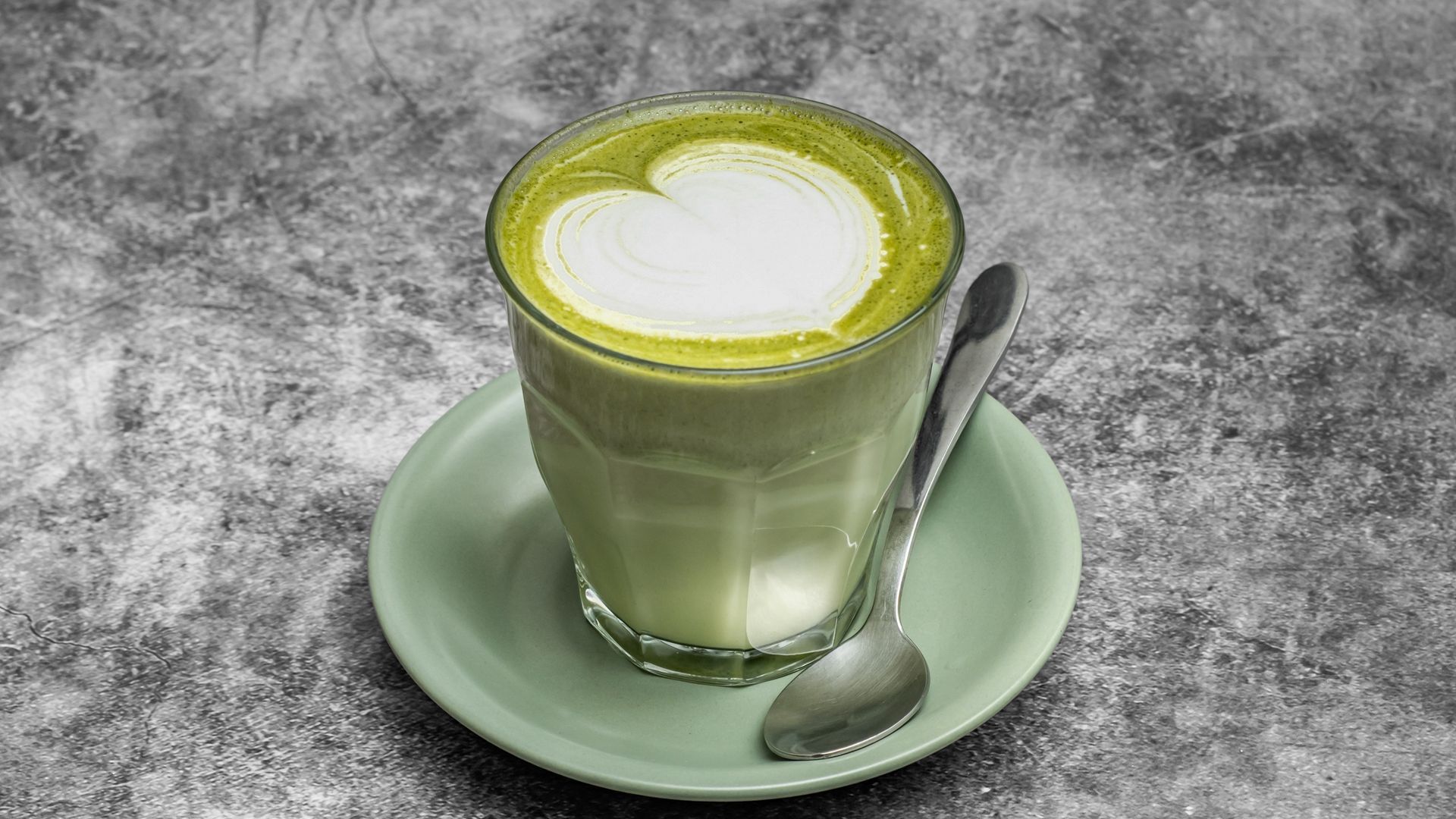 Matcha latte in a glass with a heart design, on a green saucer, with a spoon, on a gray surface.