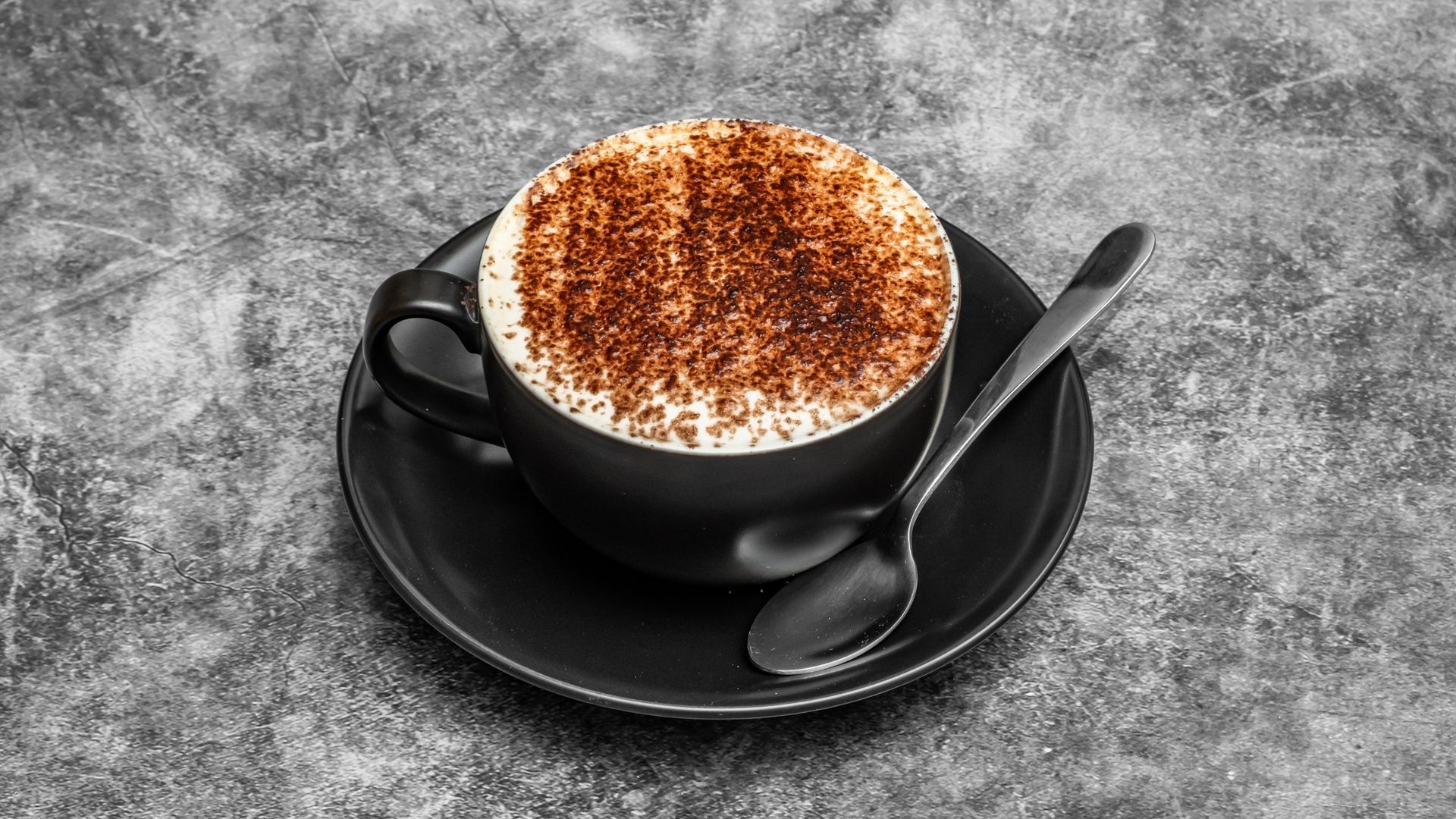 Cappuccino in a black cup with cocoa powder, on a black saucer with a spoon, on a textured gray surface.