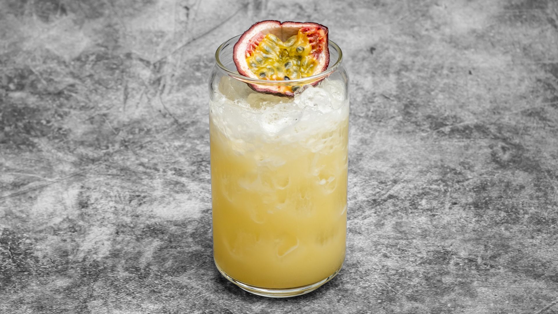 Tall cocktail in a glass, yellow liquid with crushed ice, garnished with a passion fruit slice. Gray background.
