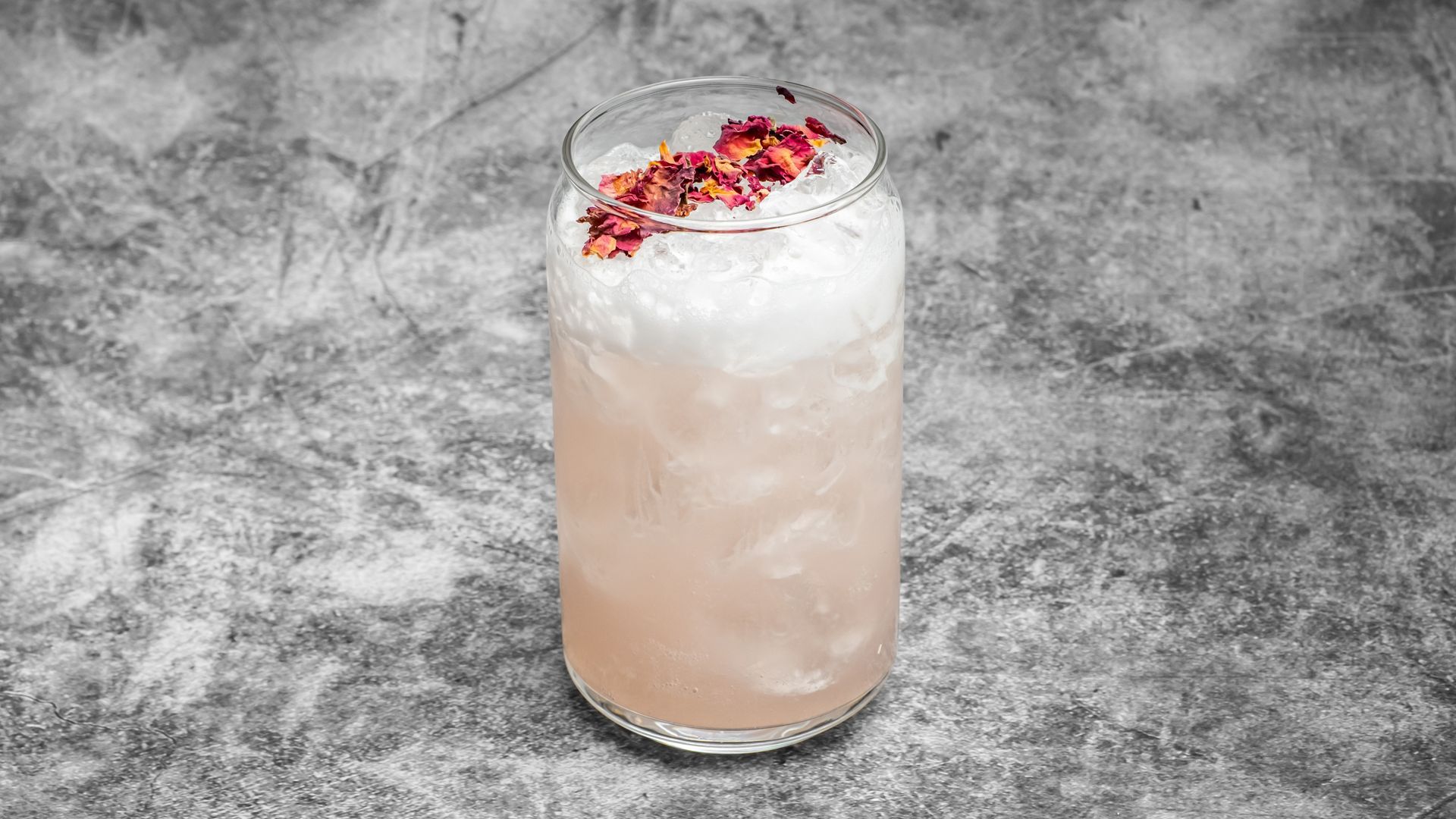 Tall cocktail in a clear glass, pale pink, topped with foam and dried rose petals.
