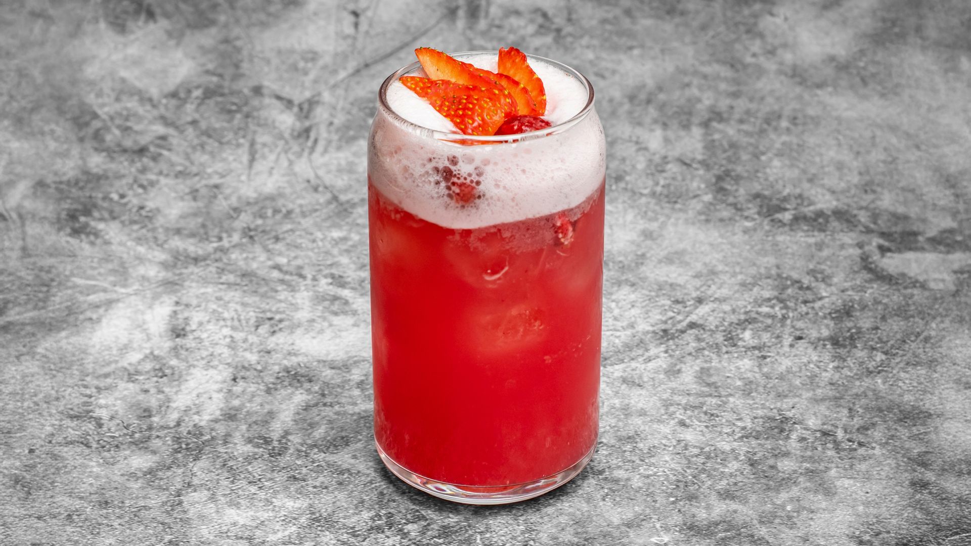 Red cocktail with white foam, garnished with strawberry slices, in a clear glass.