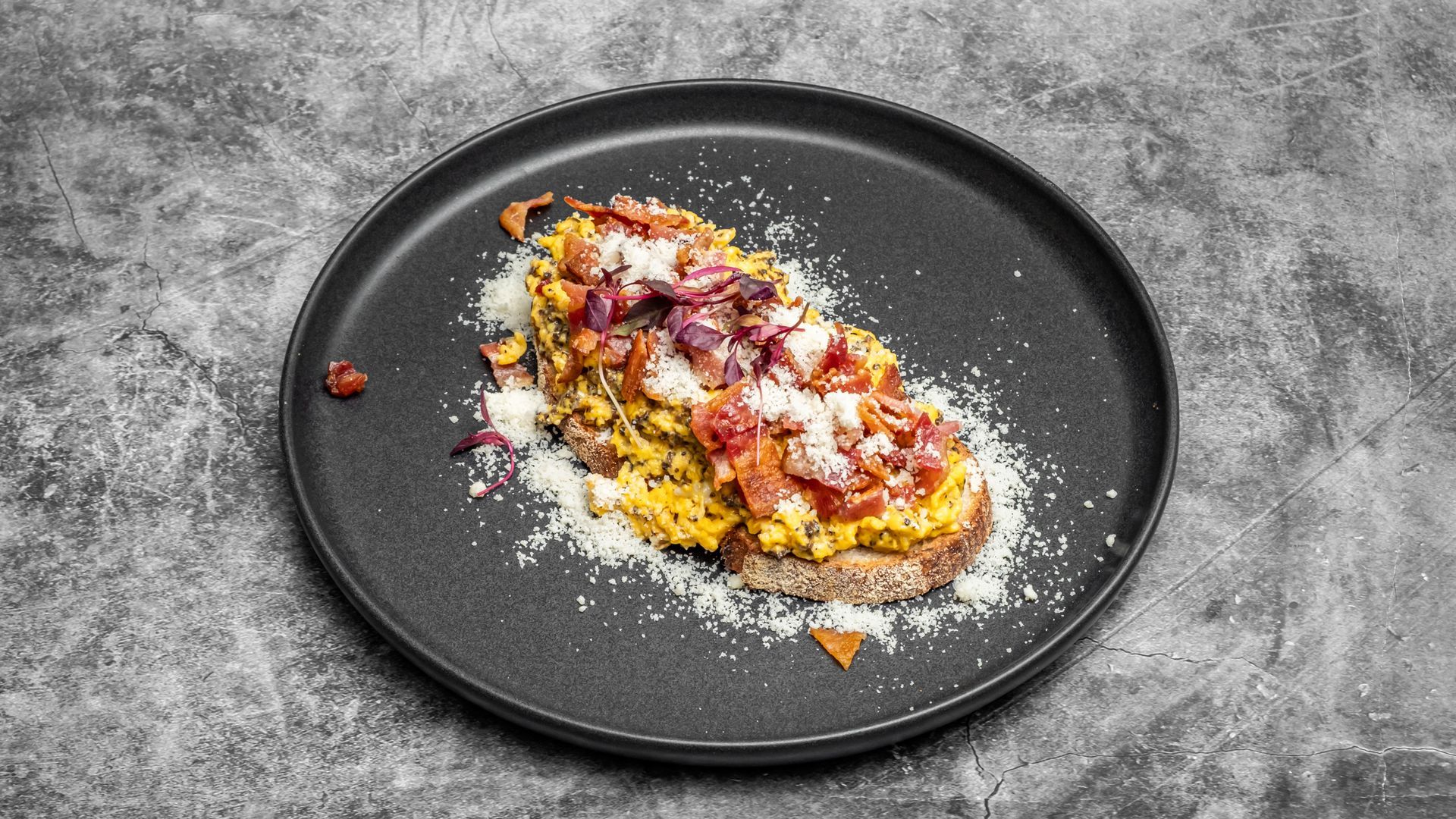 Scrambled eggs with toppings on toast, served on a black plate against a gray background.