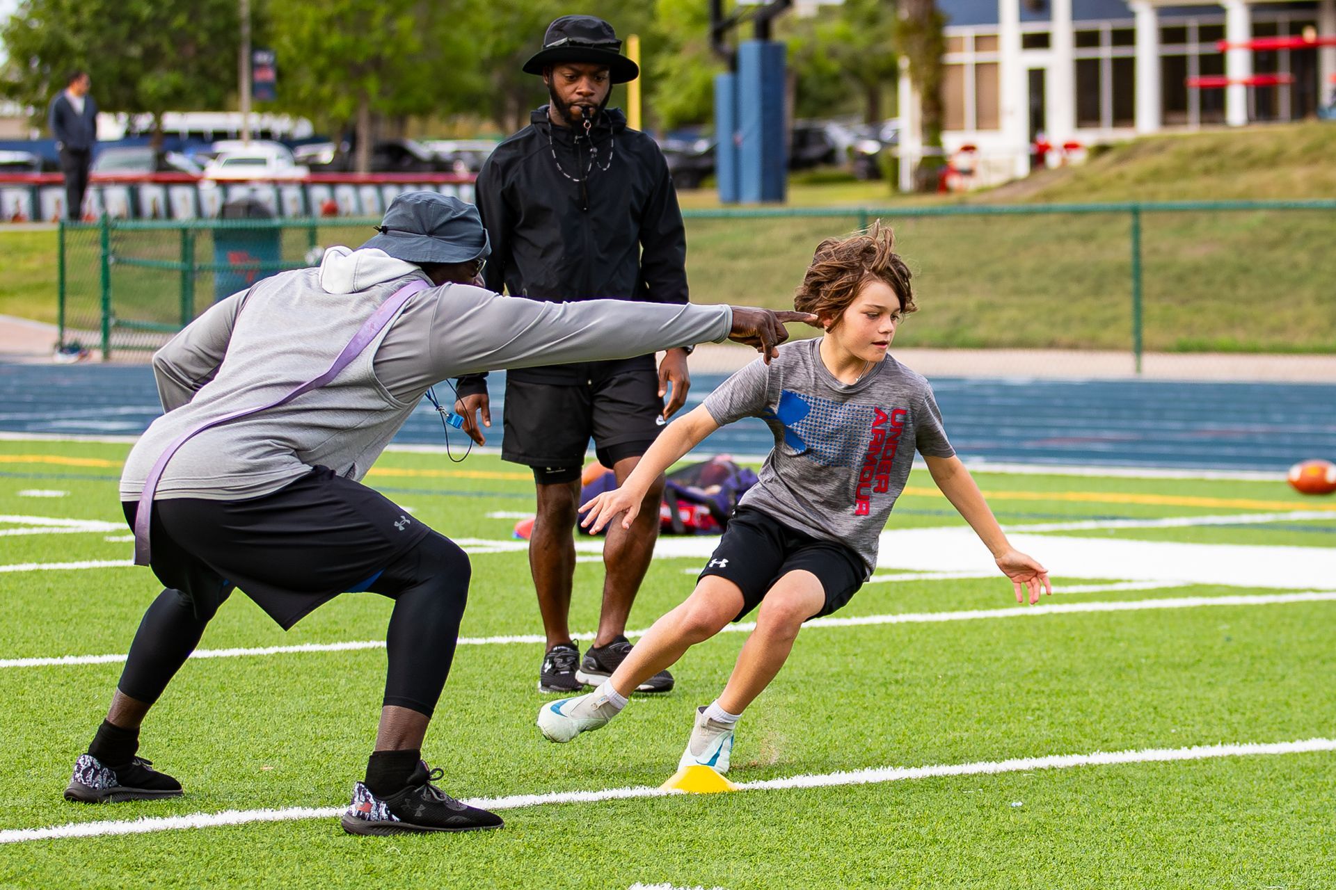 A coach in a gray hoodie gestures to a child running through an agility drill on a green football field.