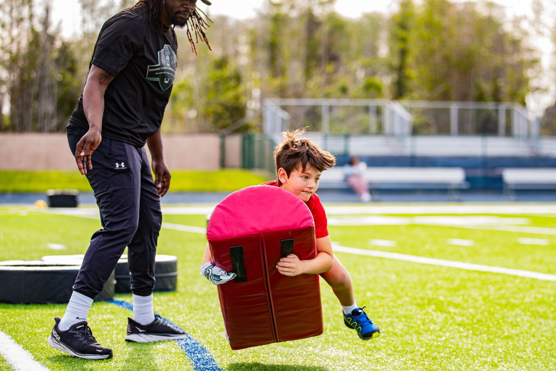 An empty football field with green artificial turf, white yard lines, and stadium seating under a cloudy sky.