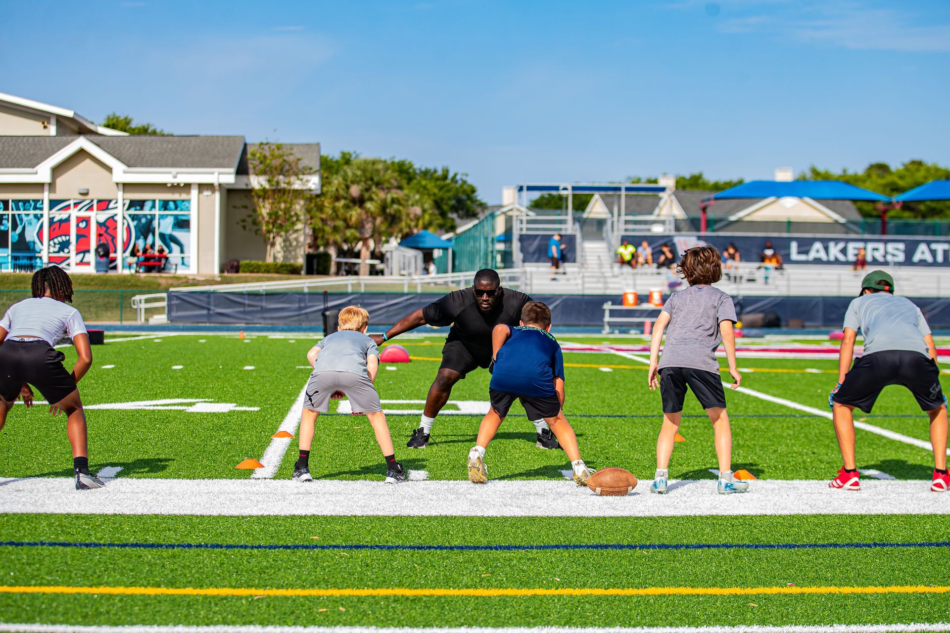 A coach stands on a field at night, holding a clipboard and speaking to a group of football players in black uniforms.