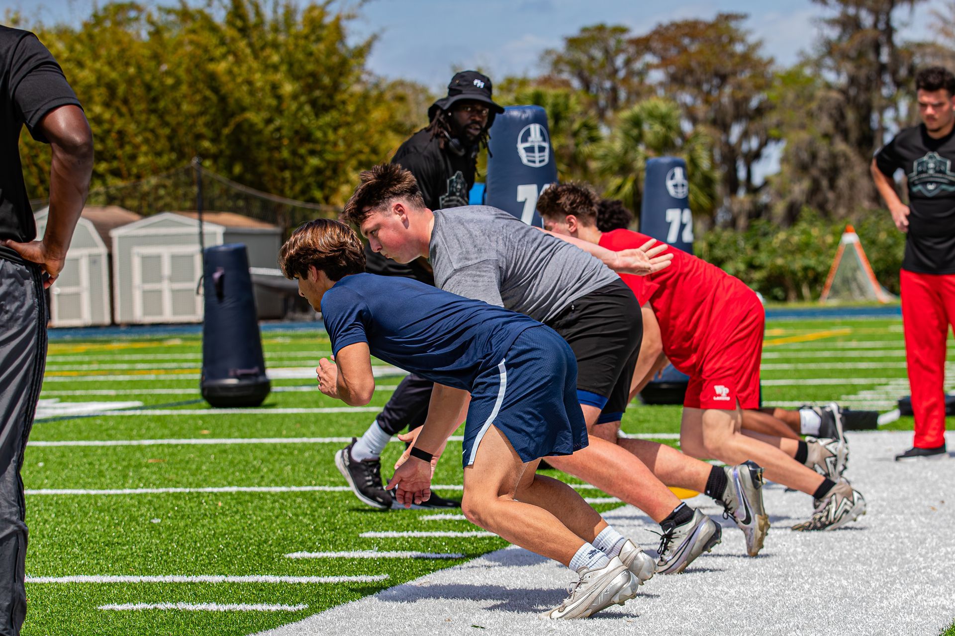 Athletes in a three-point stance during a practice drill on a sunny football field, coached by a person in a bucket hat.