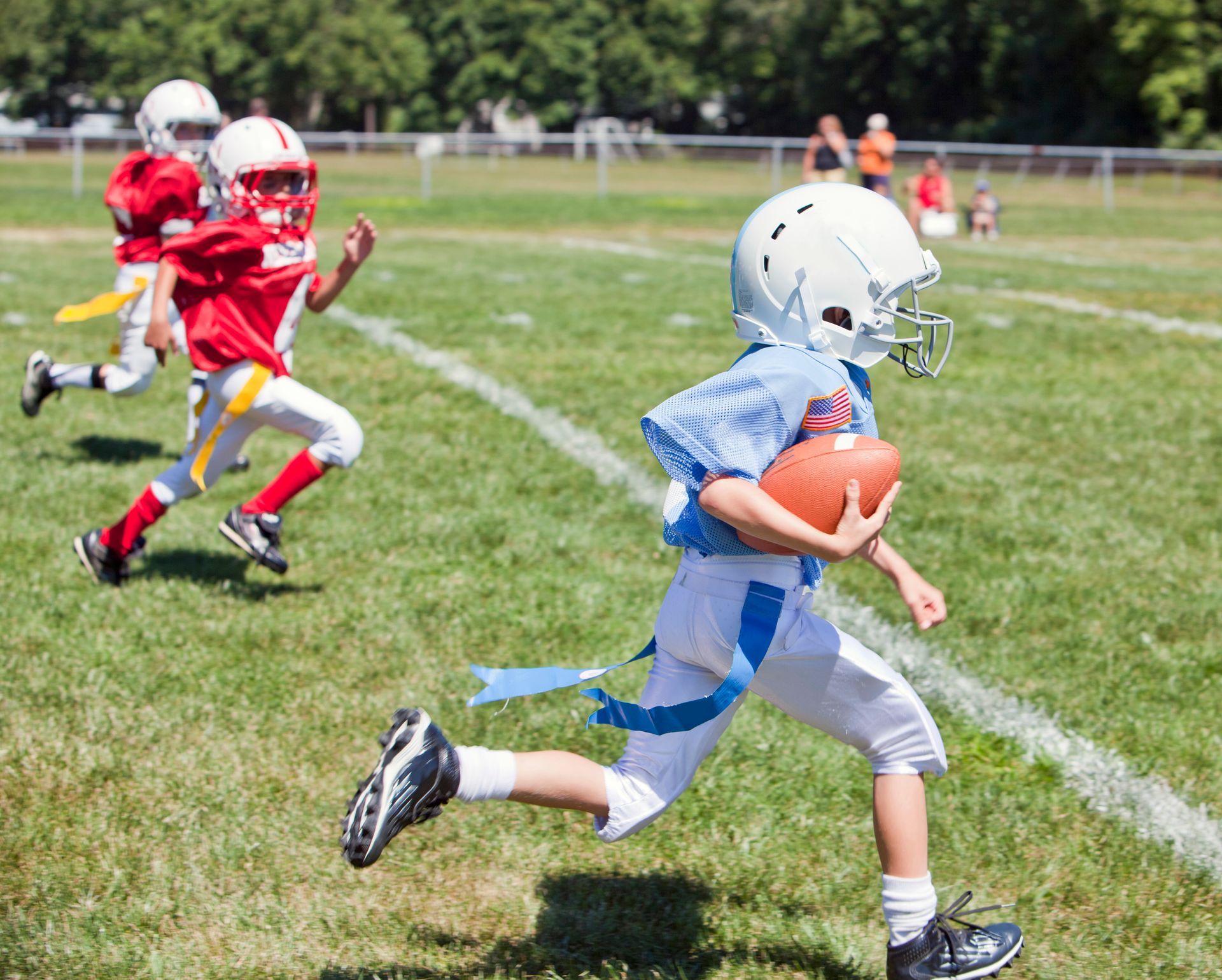 Three children in uniforms playing flag football on a grassy field, with one runner in blue carrying the ball.