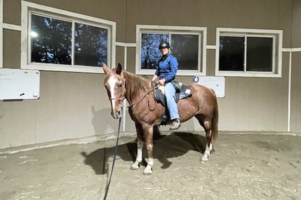 Two riders on horses, decorated with orange and green, outside a barn. Sunny day.