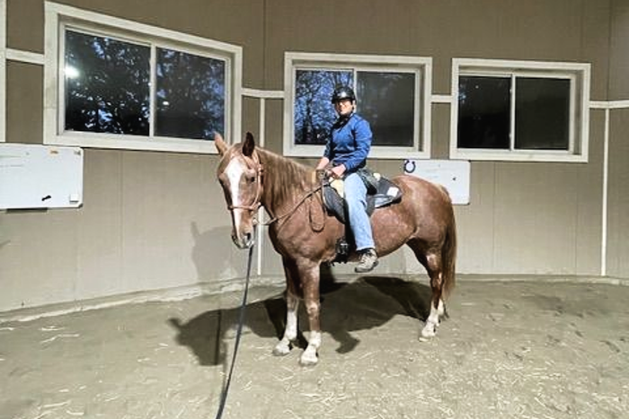 Two riders on horses, decorated with orange and green, outside a barn. Sunny day.