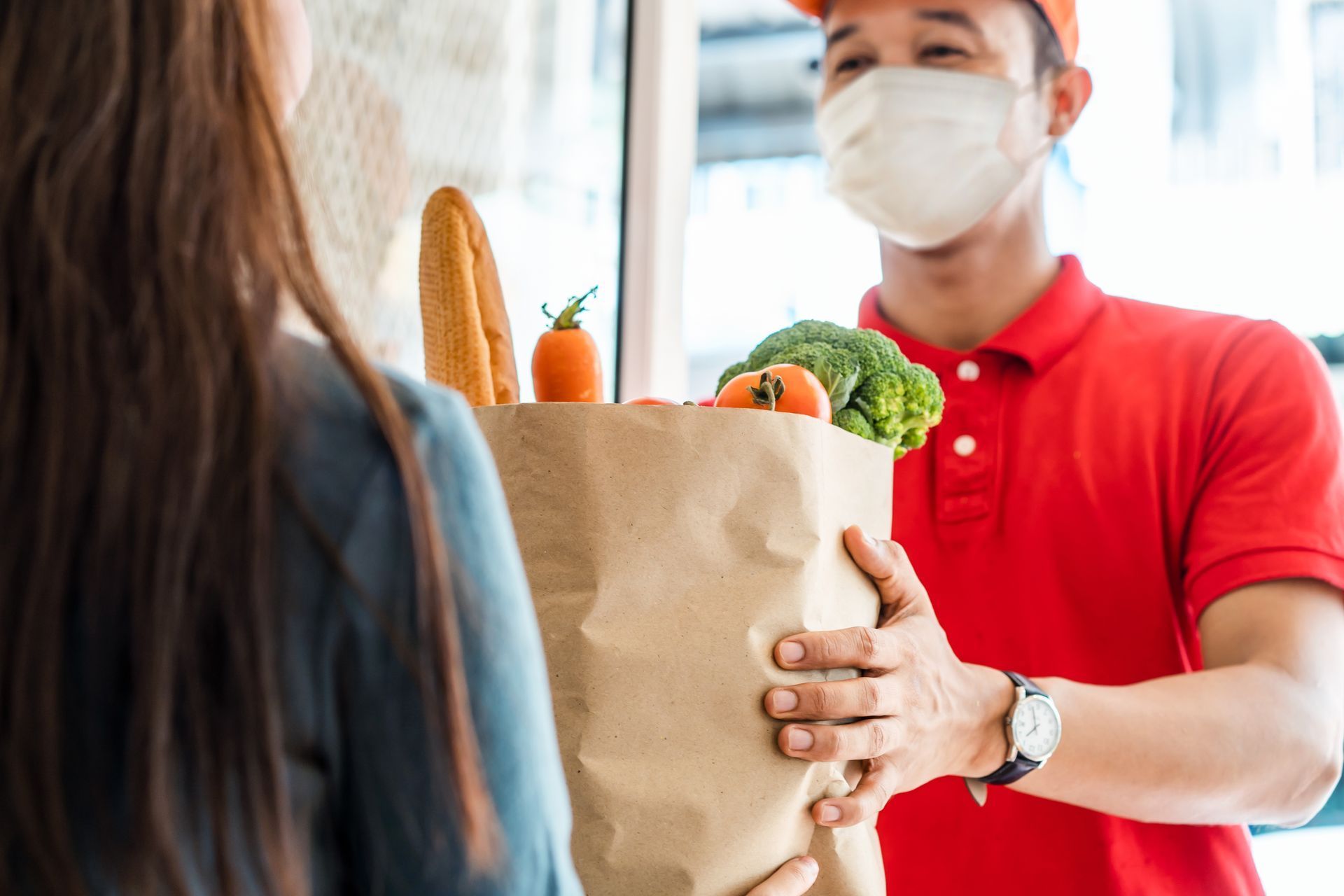 Delivery person in red shirt and mask handing a brown paper grocery bag to a person at a doorway.