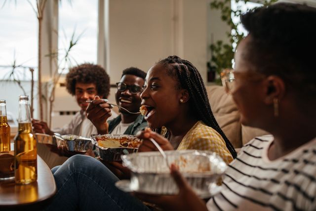 Four people seated, eating from takeout containers in a living room. They are smiling. Beverages on table.