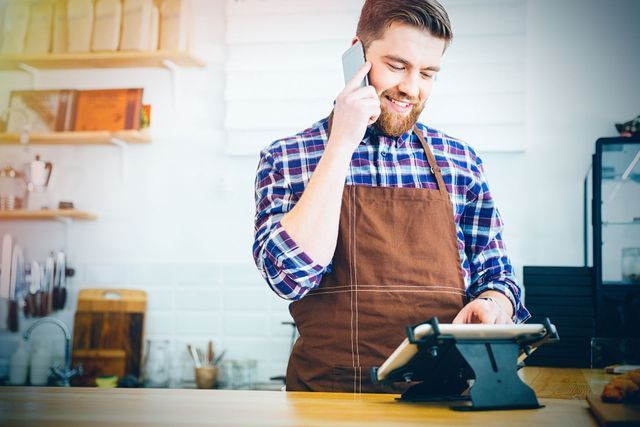 Barista in brown apron smiles while talking on phone and using tablet. Counter setting.
