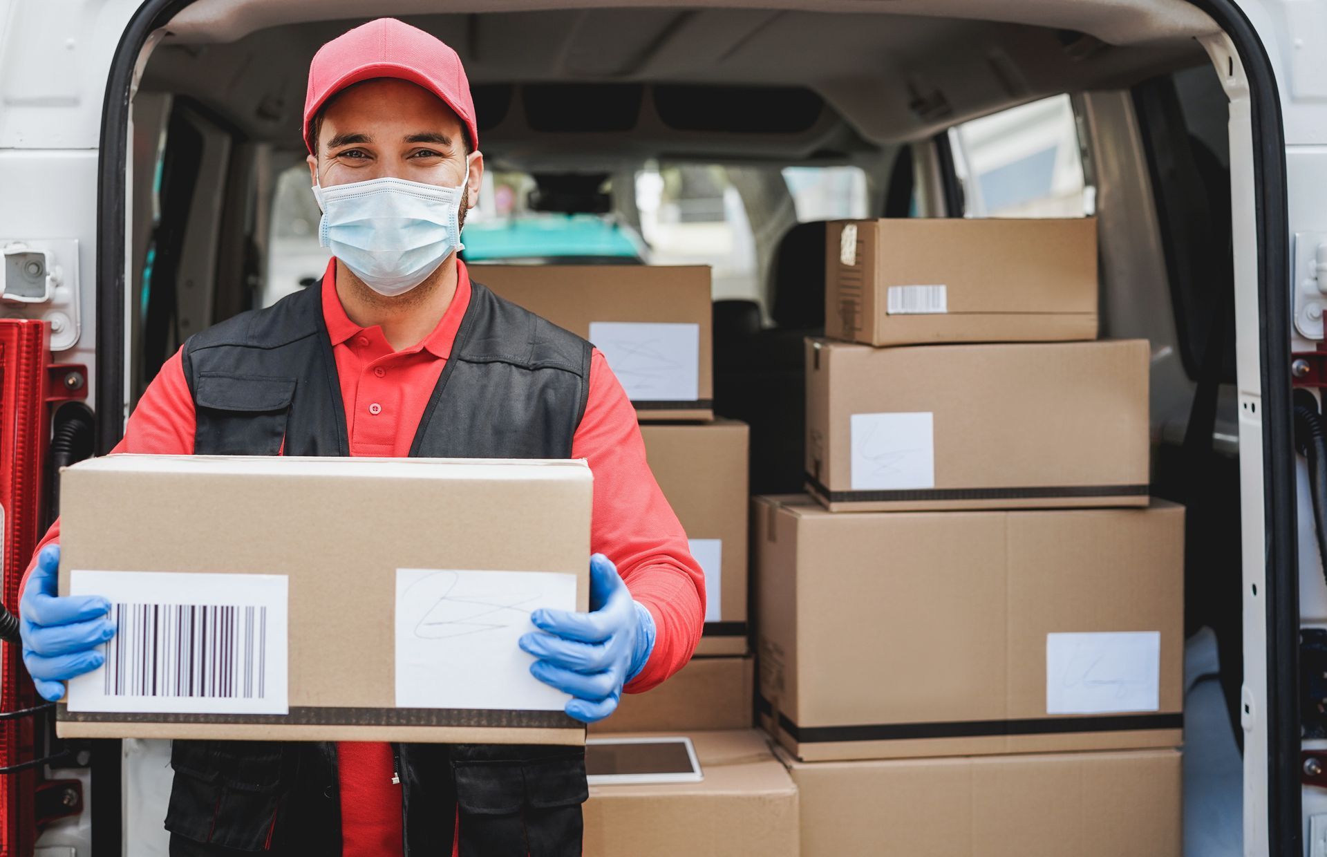 Delivery person in mask, gloves, holding package, surrounded by boxes in a van.