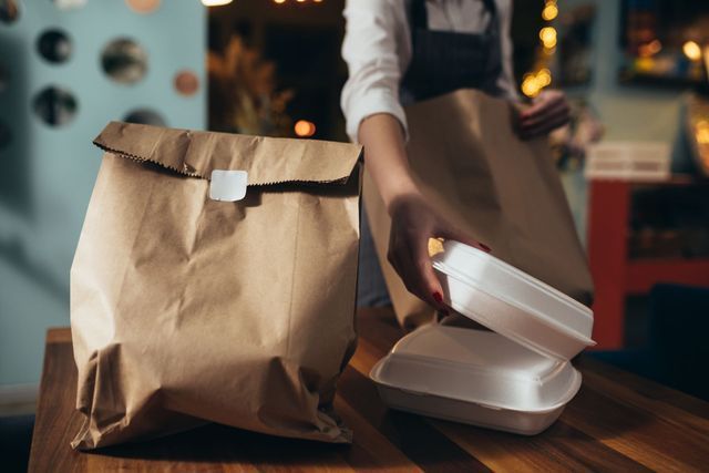 Person packing takeout containers into a brown paper bag on a wooden table.