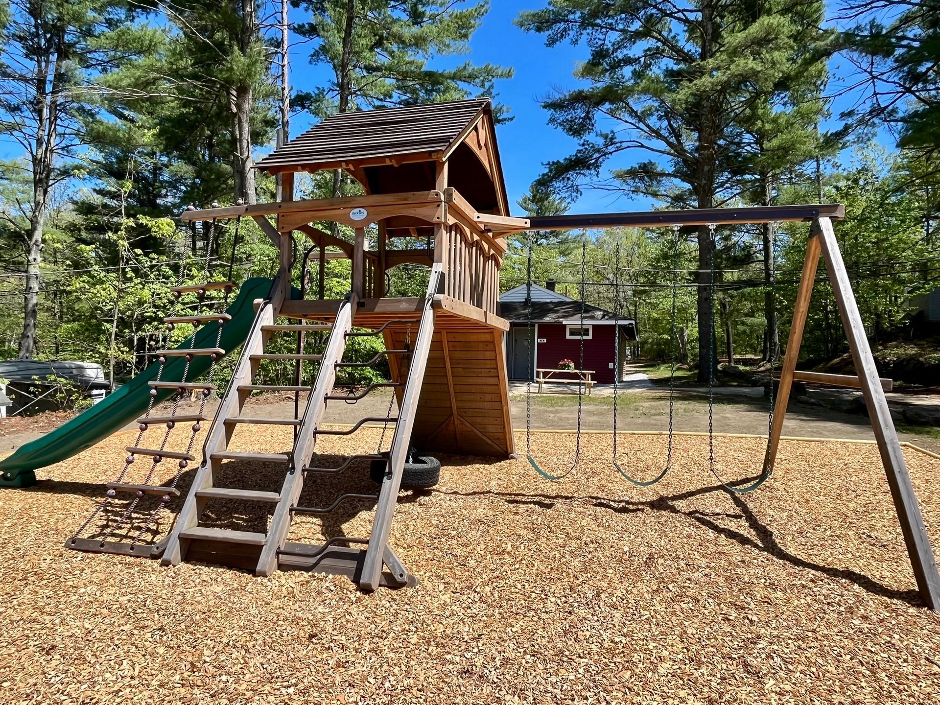 Wooden playground with slides, climbing steps, and swing set on a woodchip surface among pine trees.