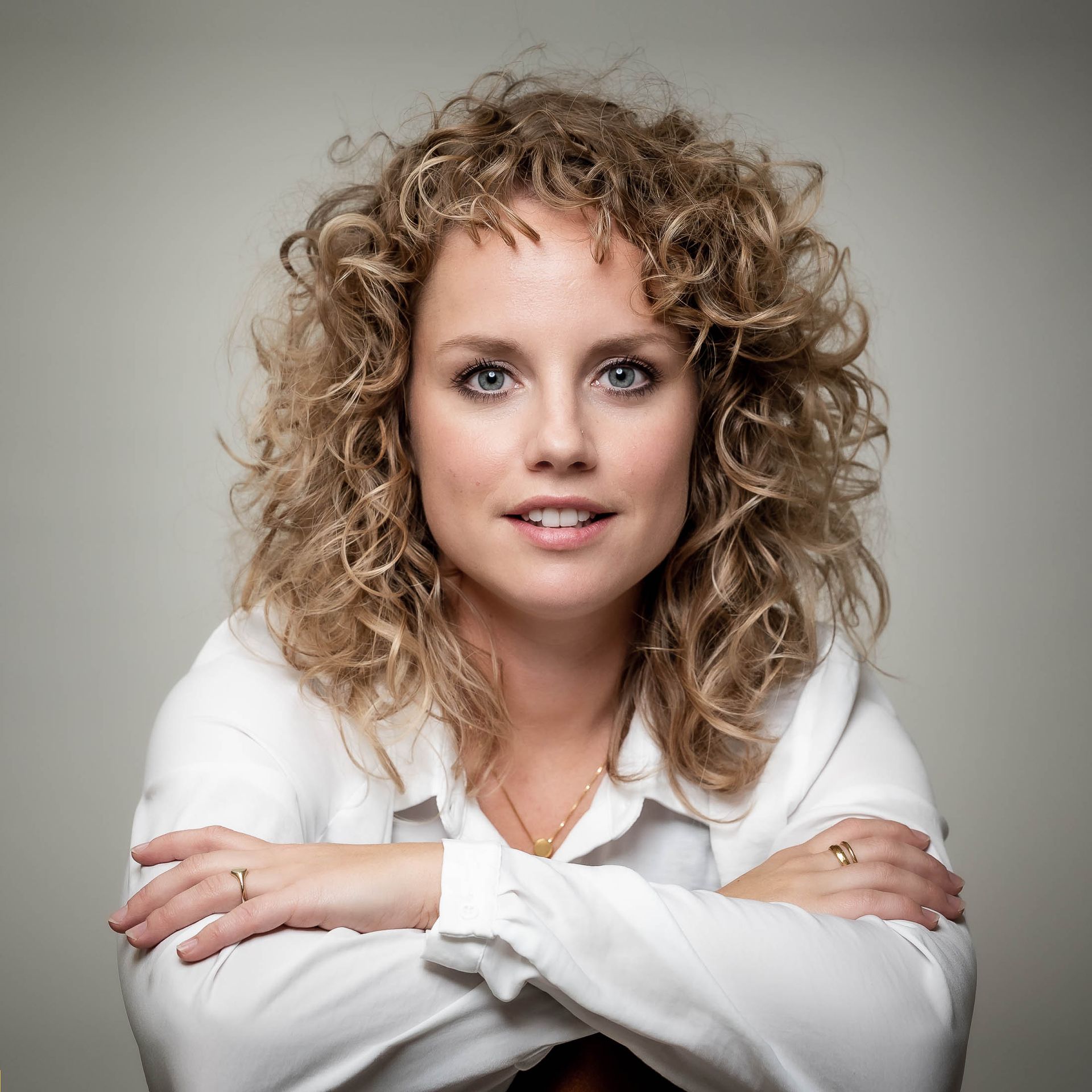 Een vrouw met krullend haar draagt een wit shirt en heeft haar armen over elkaar geslagen.