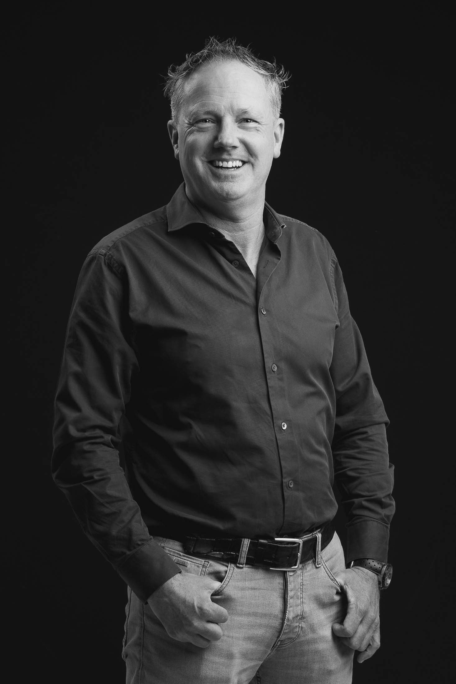 Een man in een zwart shirt en spijkerbroek lacht op een zwart-witfoto.