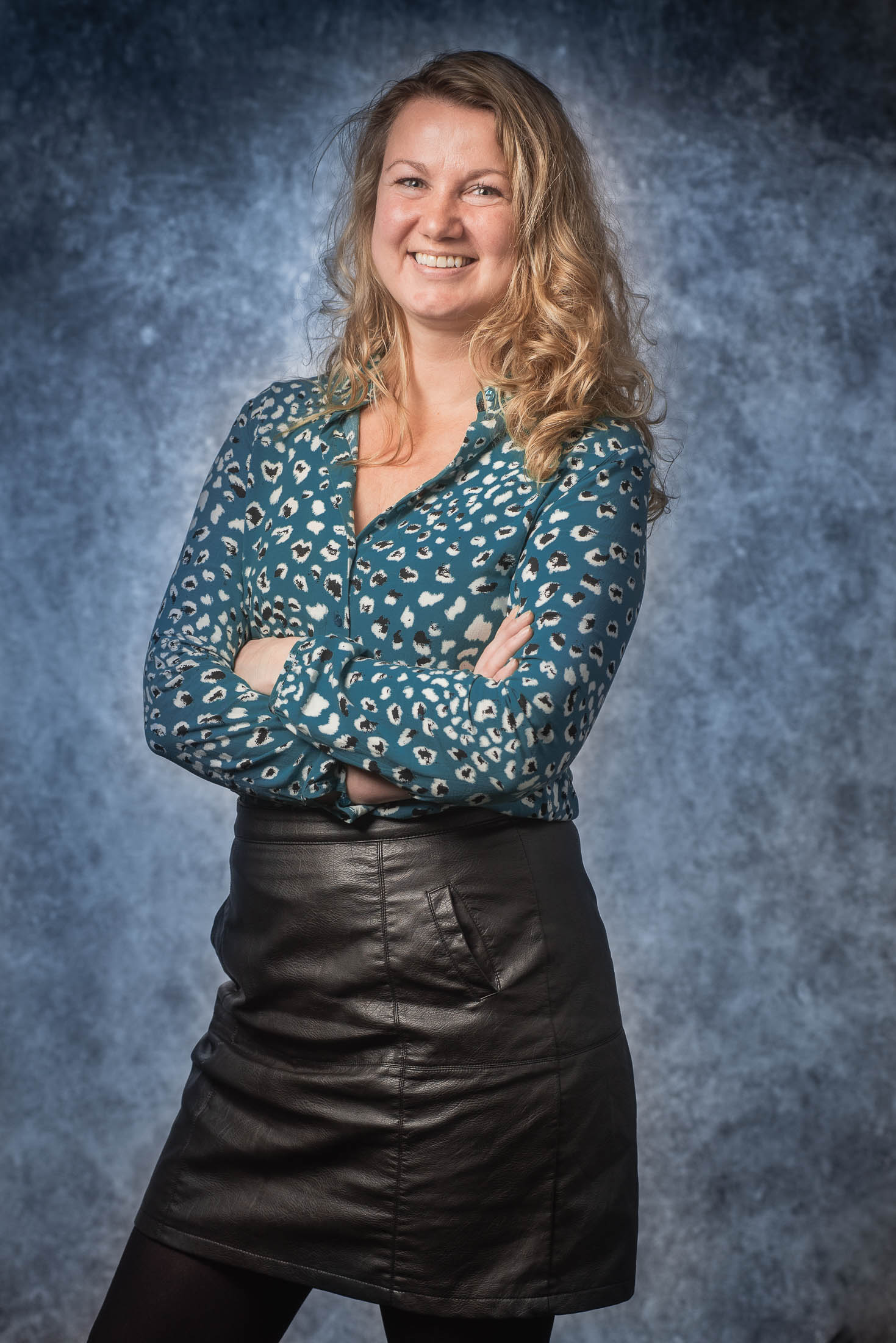 Een vrouw in een blauw shirt en een zwarte rok staat met haar armen over elkaar.