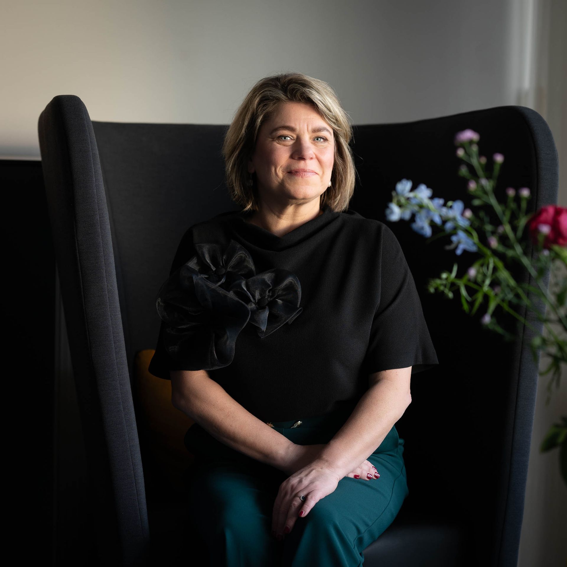 Vrouw in zwarte top en groene broek, zittend op een zwarte stoel met hoge rugleuning, kijkt opzij. Bloemen op de achtergrond.