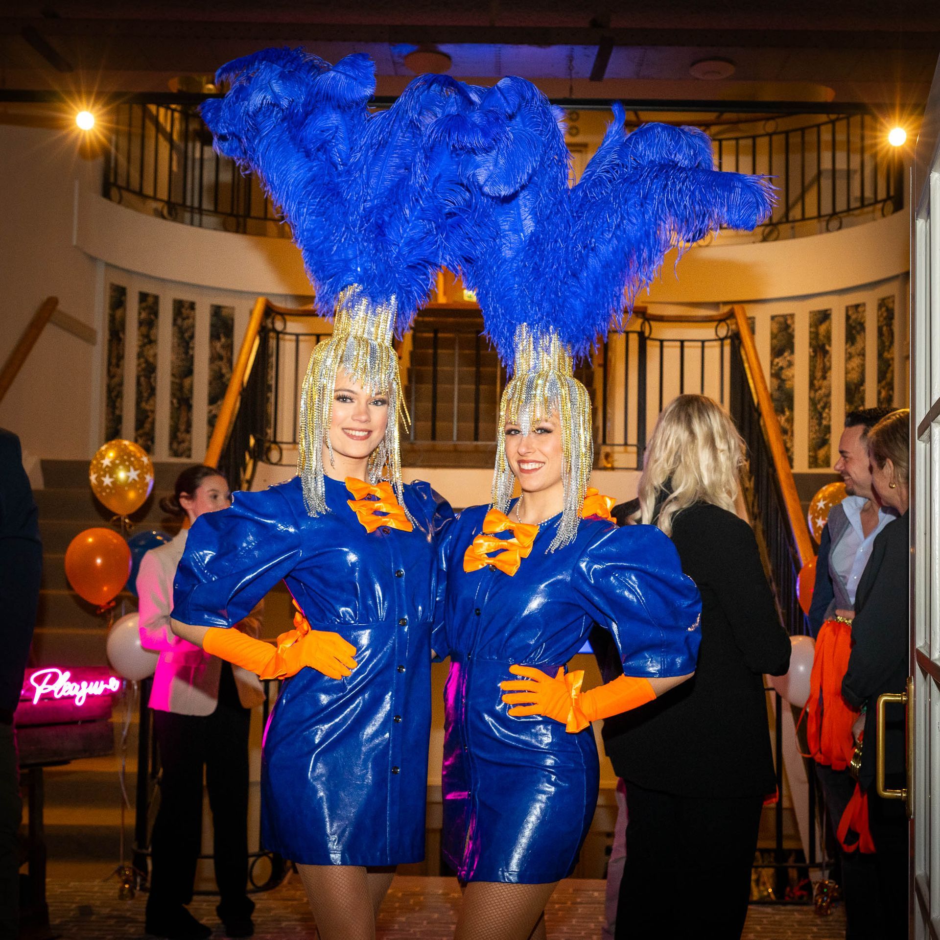 Twee vrouwen in glanzende blauwe jurken en hoofdtooien met veren poseren bij een trap tijdens een evenement.