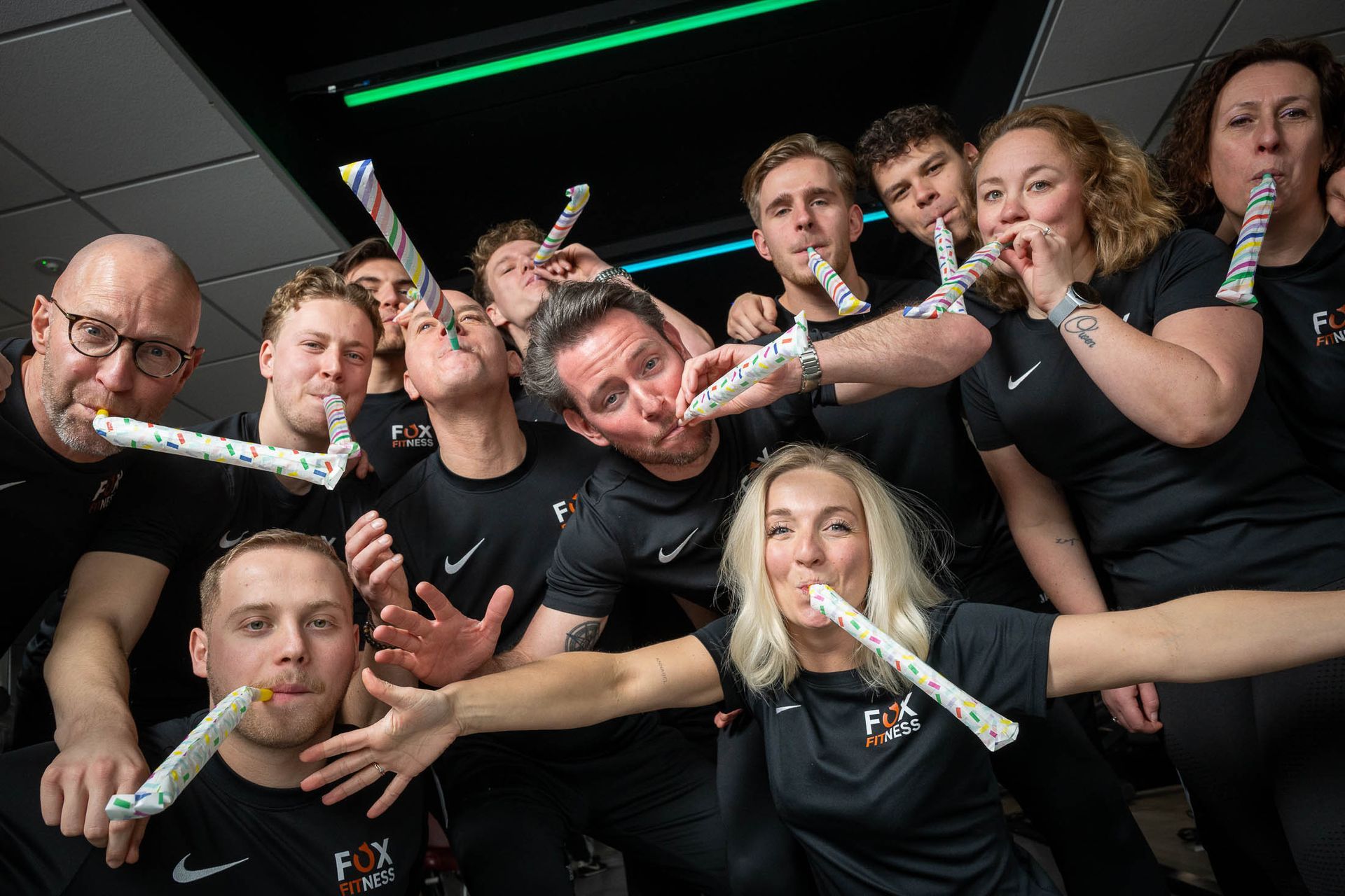 Een groep mensen die op feesttoeters blazen, poseren in een donkere ruimte met groen licht. Ze dragen zwarte shirts en glimlachen.