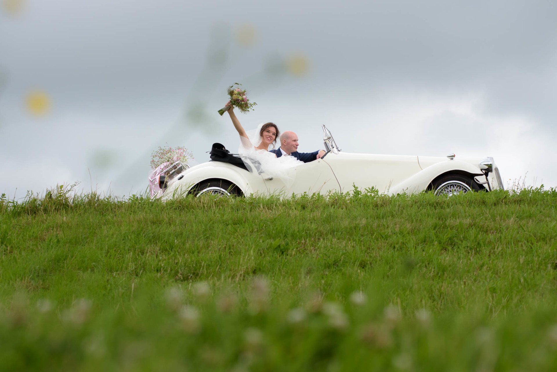 Een bruid en bruidegom rijden in een witte auto op de top van een grasheuvel.