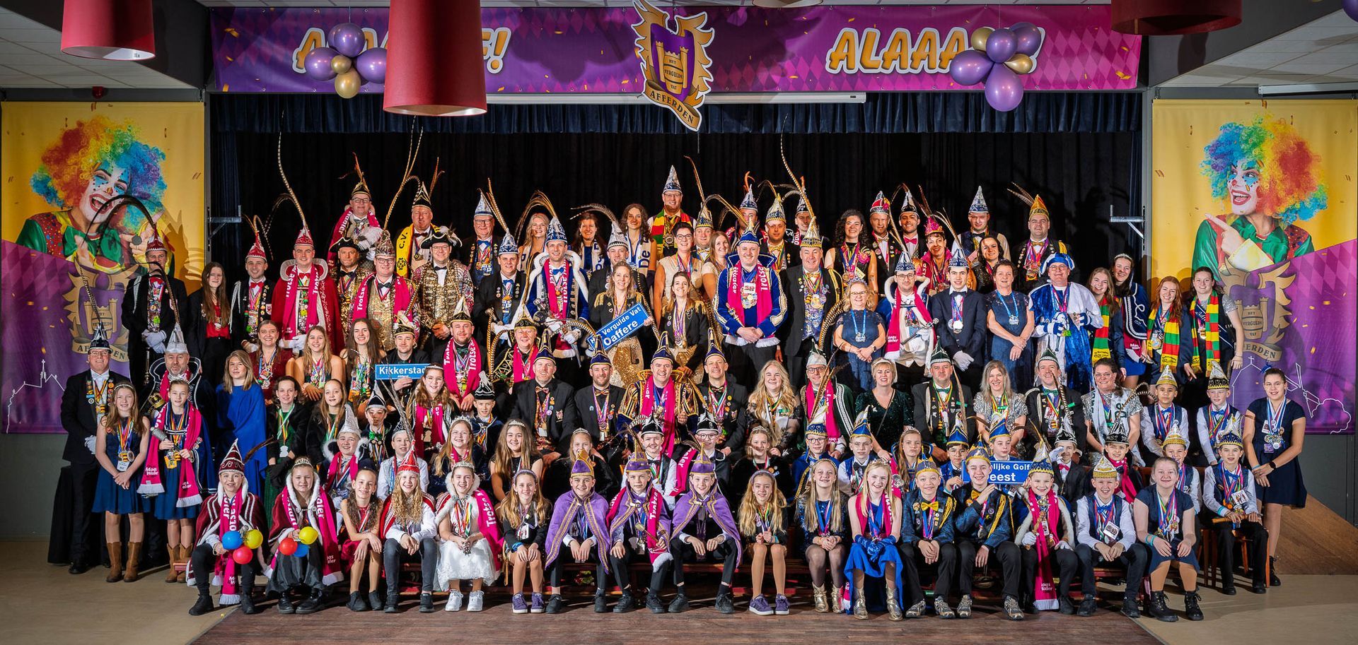 Een grote groep mensen in kostuums poseert op een podium, mogelijk voor een carnaval of feest, in een versierde zaal.