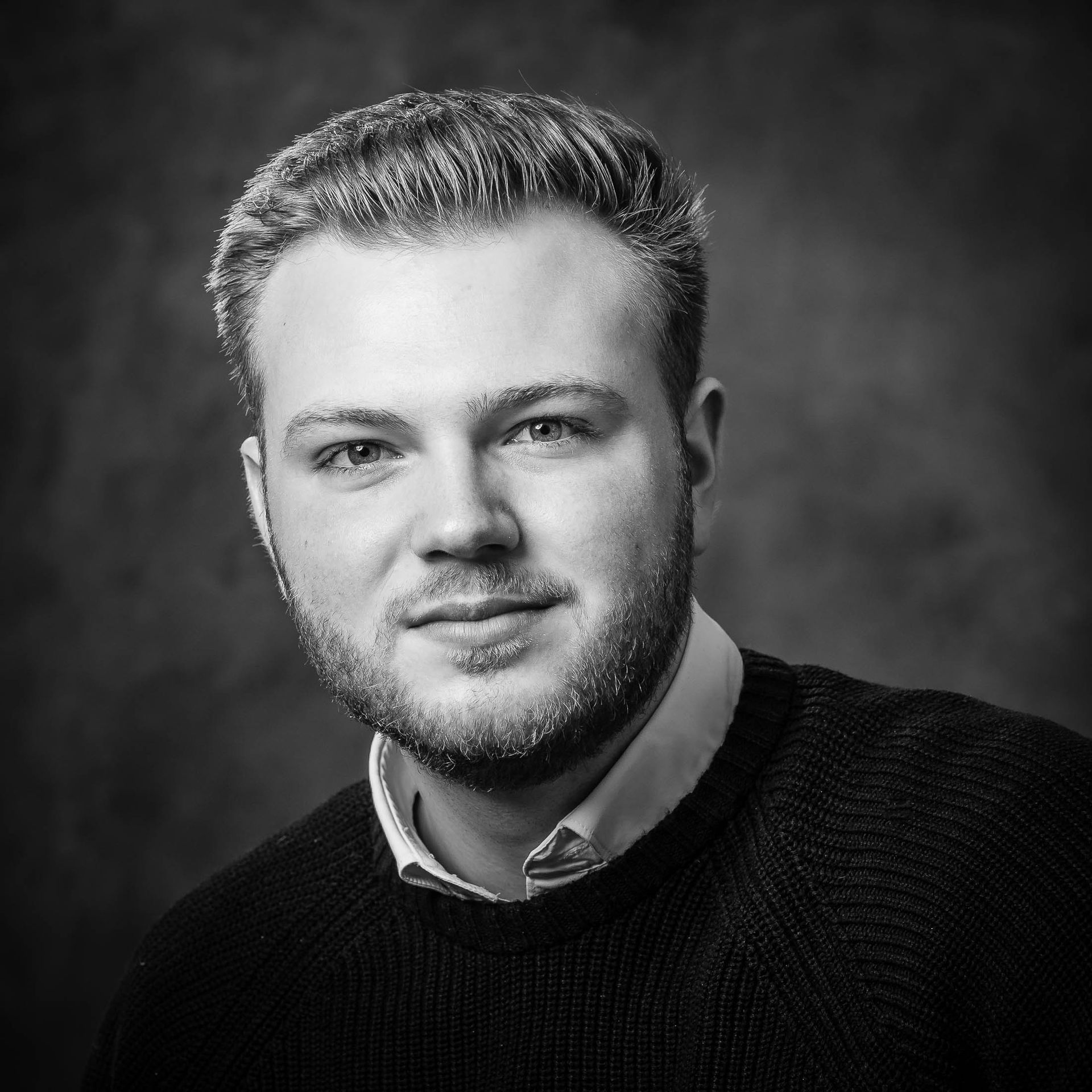 Een man met een baard draagt een zwarte trui en een wit overhemd.