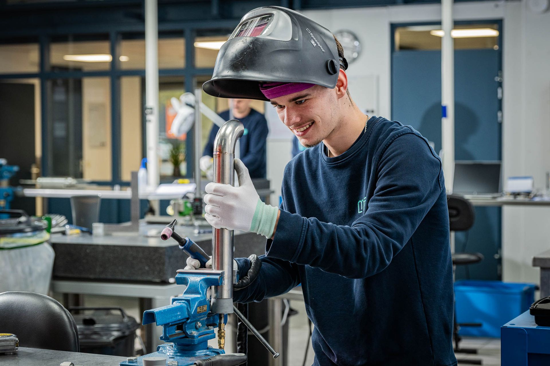 Een lasser in een werkplaats, lachend terwijl hij met metaal en gereedschap werkt.